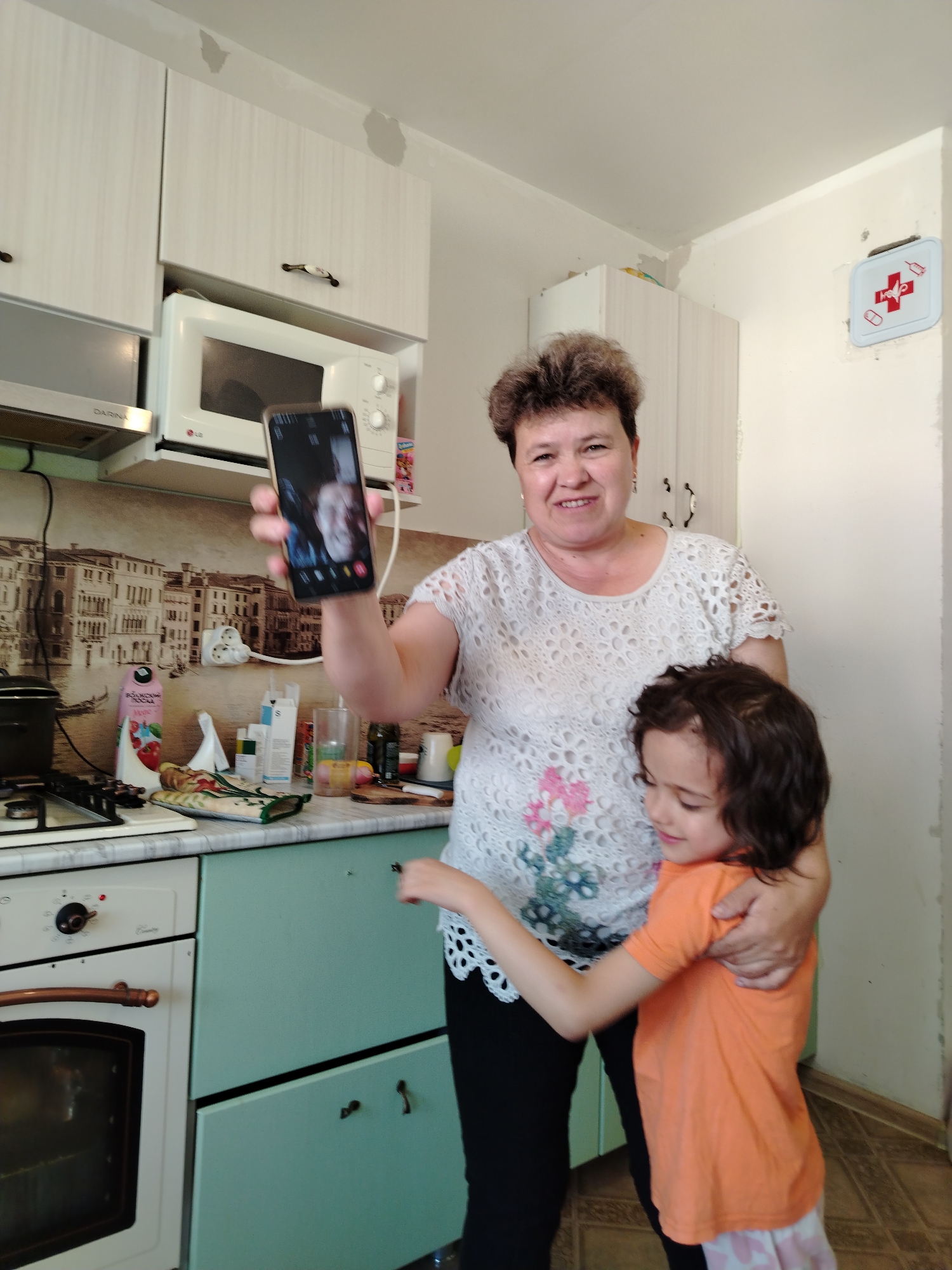 The image shows a woman and a young girl in a kitchen. The woman is holding up a smartphone, appearing to be taking a selfie or participating in a video call. The girl is standing in front of her, being embraced by the woman. Both subjects are smiling, suggesting a positive and happy interaction. The kitchen appears to be modestly equipped, with cabinets, a microwave, a stove, and various kitchen items on the countertops. There is a patterned backsplash behind the counter, and a first-aid kit is mounted on the wall. The lighting suggests it is daytime. There is no visible text in the image.
