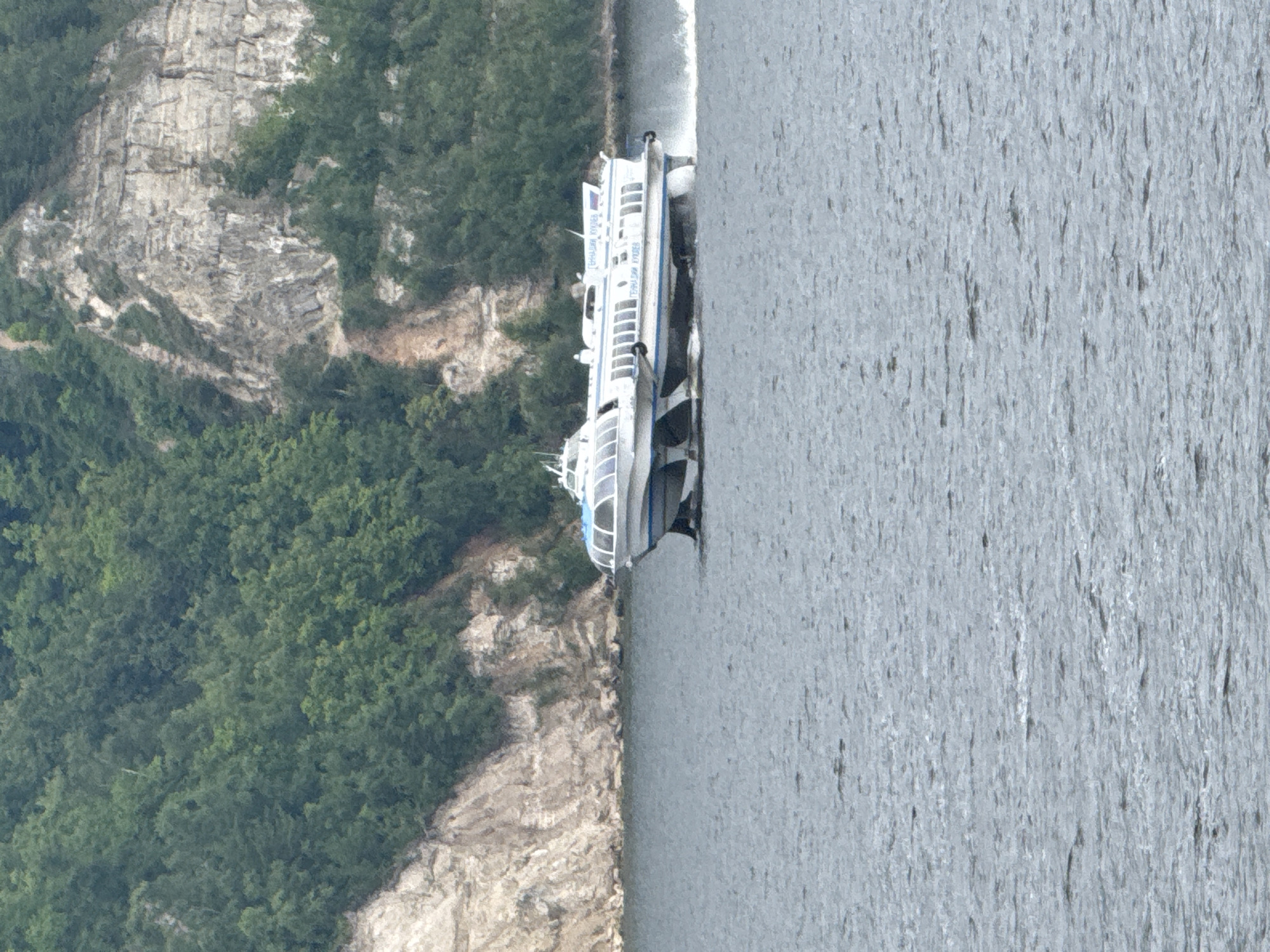 A white hydrofoil vessel with blue trim and a glass-enclosed cabin is moving at speed on a gray body of water. The vessel has foil-like structures beneath it, which are partially submerged and creating a wake. In the background, a steep, rocky cliff face covered in lush green trees rises from the water's edge. The sky is not visible in the frame. The text "ГЕЙНАДИЙ БУХОВ" is visible on the side of the vessel. The location is Kamskoye Ustye, Russia.