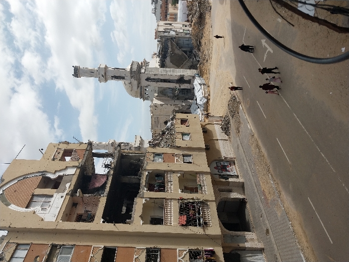 The image captures a scene of extensive structural damage in Khan Yunis, Palestinian Territory, under a partly cloudy sky. A multi-story residential building dominates the left, exhibiting severe damage with collapsed sections, exposed rebar, and large openings revealing interiors. Its light-colored facade is marked by numerous impact points, and some balconies still show hanging fabrics. Arched architectural details are partially destroyed on upper floors. At ground level, arched entrances and graffiti are visible, with small piles of debris accumulating near the building's base and along the sidewalk.
In the midground, to the right of the damaged building, stands a significantly damaged mosque, identifiable by its white minaret and dome, both showing structural compromise. Extensive piles of rubble and debris surround the mosque and extend into the background, where additional destroyed or damaged structures are visible.
A paved road runs horizontally across the lower frame, featuring white lane markings, including a turn arrow. A patterned sidewalk parallels the damaged building. On the road, a group of five individuals, comprising two adults and three children, walks from left to right. Behind them, one person pushes a stroller or wheelchair. Further down the road, two solitary figures are visible; one walks on the right side, and another is in the distant rubble. Small figures are also discernible within the damaged upper floors of the residential building. A black cable partially obscures the lower right foreground.