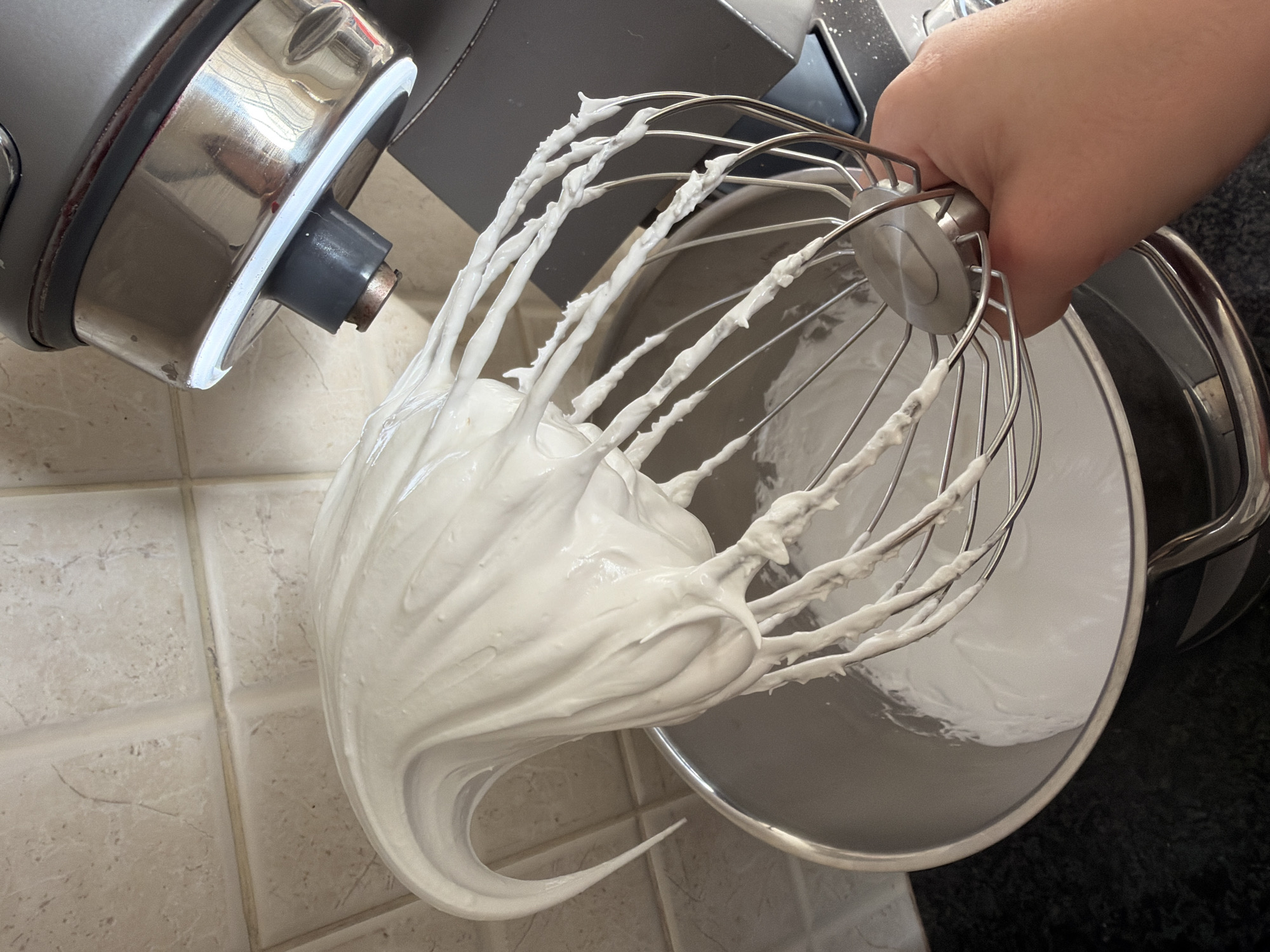 The image displays a close-up view of a culinary activity taking place in a kitchen. The main subject is a metal whisk attachment, held by a human hand in the upper right portion of the frame. The whisk is heavily coated in a thick, white, aerated mixture, which appears to be either meringue or whipped cream. This mixture exhibits stiff peaks and a distinctive, elongated curve, indicating a well-whipped consistency.
Below the whisk, a shiny, stainless steel mixing bowl, also containing a quantity of the white mixture, is visible. To the left, a portion of a stand mixer's head unit, featuring metallic gray and silver components, suggests the equipment used for the preparation. The setting is clearly an indoor kitchen, characterized by light-colored, possibly beige or off-white, square tiles on a surface that could be a wall or backsplash. A darker countertop surface is discernible in the background to the right.
The image captures the moment after the whisking process, highlighting the texture and stability of the prepared mixture. The lighting is bright and even, emphasizing the clean, white appearance of the food. No visible text is present in the image. This scene is set in Tel-Aviv, Israel.