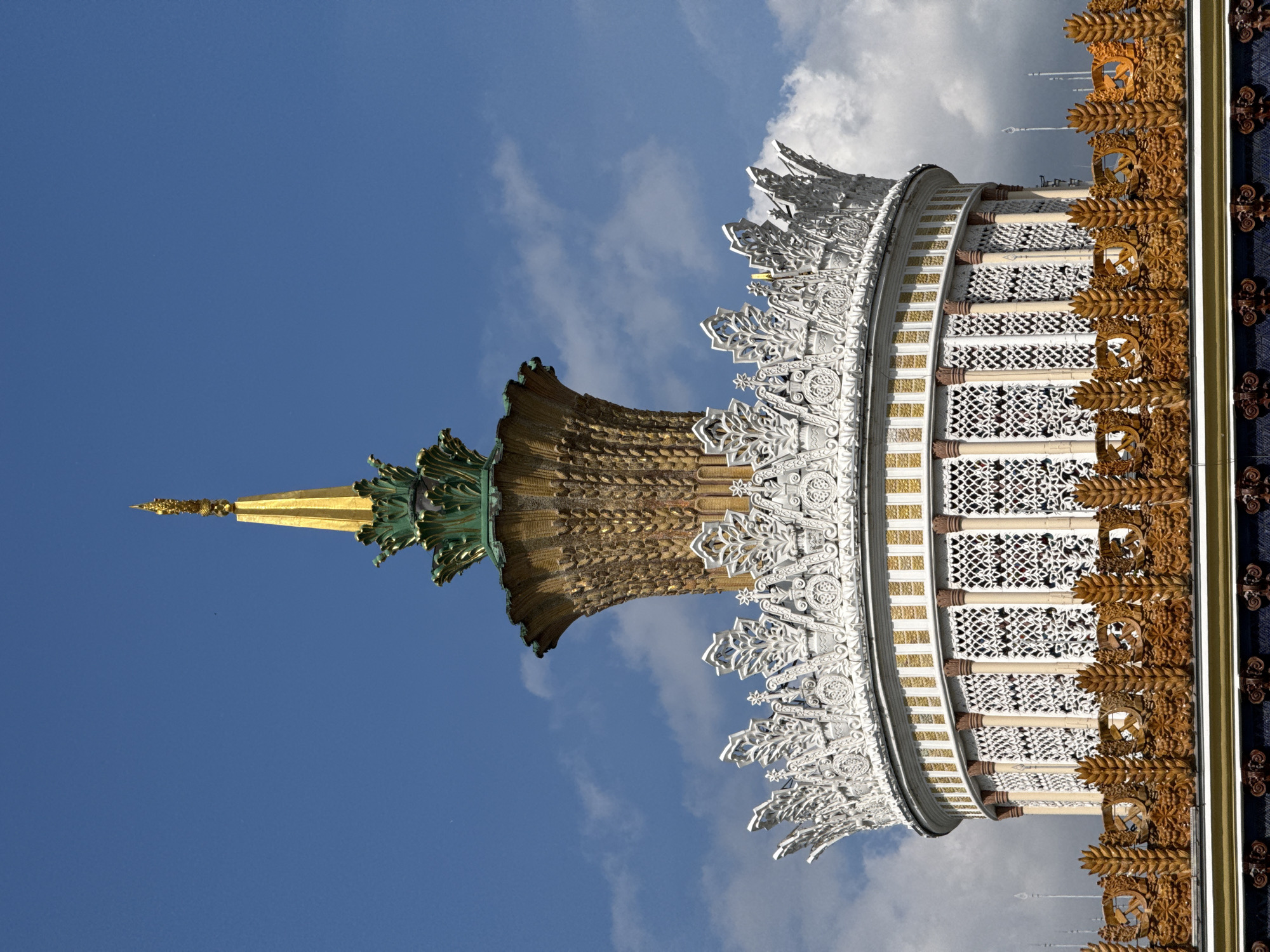 The image depicts a portion of the Moscow State University main building in Moscow, Russia, angled upwards against a blue sky with scattered white clouds. The architectural details include a golden spire with green accents, elaborate white and gold patterned facades, and a border featuring brown ornamentation on a dark blue background. The composition emphasizes the intricate design and scale of the building.