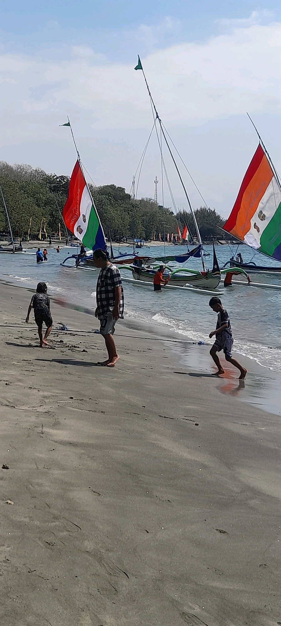 The image depicts a beach scene in Pasir Putih, Indonesia, with several traditional Indonesian fishing boats moored in shallow water near the shore. The boats have colorful sails, predominantly in shades of red, white, orange, and green, some bearing emblems. Two young boys and a man are present on the sandy beach. One boy, wearing a dark patterned shirt and shorts, is walking away from the camera towards the water. The man, dressed in a checkered shirt and shorts, stands facing the water, looking towards the boats. Another young boy, in a dark patterned shirt and blue shorts, is running along the water's edge, casting a shadow. The water is calm and clear, with gentle waves lapping the shore. Trees and foliage line the background, with some structures visible in the distance, including telecommunication towers. The sky is bright and partly cloudy.