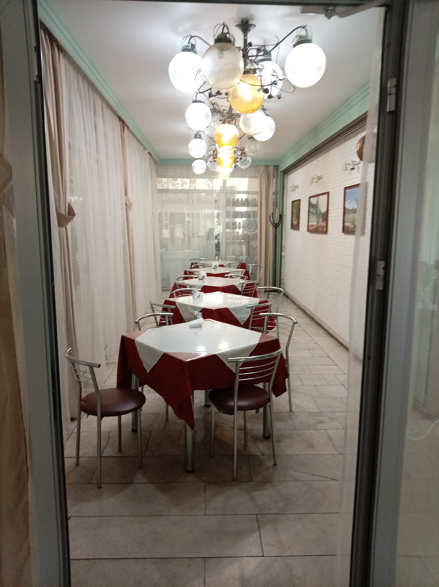 This is a medium-shot, eye-level, indoor photograph of a restaurant's dining area in Zheleznovodsk, Russia. The perspective is from just outside the entrance, looking down a long, narrow hallway. On the left, sheer beige curtains hang from the ceiling to the floor. The ceiling features a decorative molding painted light green and a multi-bulb chandelier with round, frosted glass shades.
The room is furnished with multiple tables, each covered with a red tablecloth and a white overlay. Chairs with silver metal frames and dark red padded seats are placed around the tables. The floor is tiled with large, light-colored square tiles. The walls are white and adorned with three framed pictures. The overall impression is of a clean, simple, and somewhat dated establishment. No people are visible in the frame.