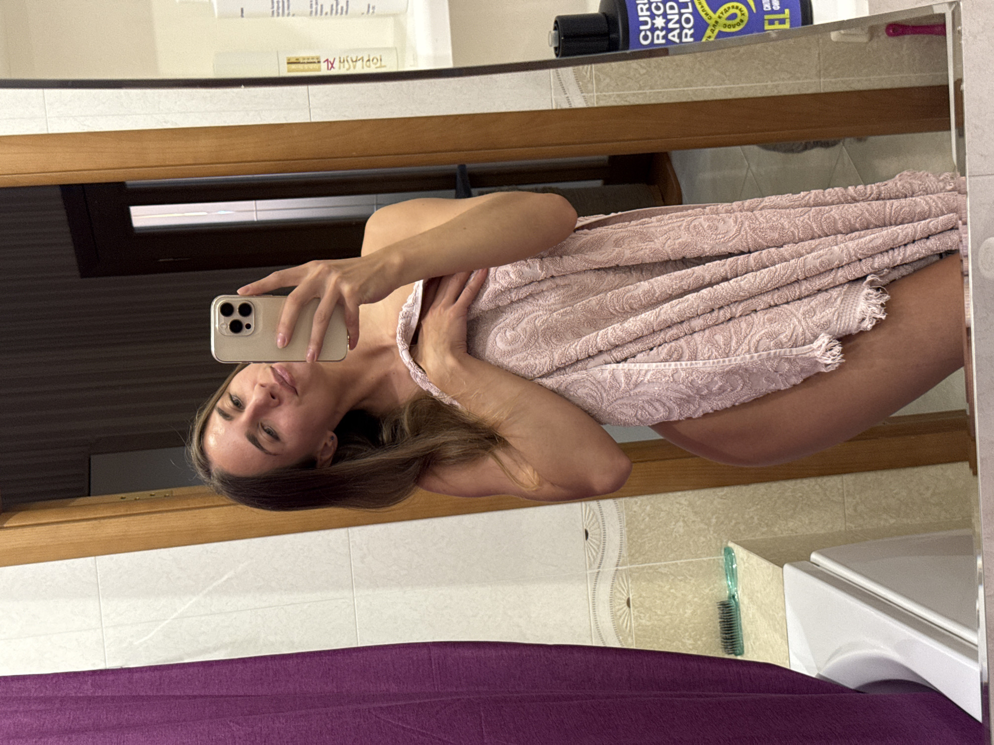 A young adult female is taking a mirror selfie in a bathroom, holding a pink towel around herself. She is wearing a gold-colored phone case. To the left of the image is a purple curtain, and to the right are shelves with various toiletries, including bottles of shampoo and conditioner. The bathroom appears to be in a residential setting, possibly an apartment in Shushary, Russia.