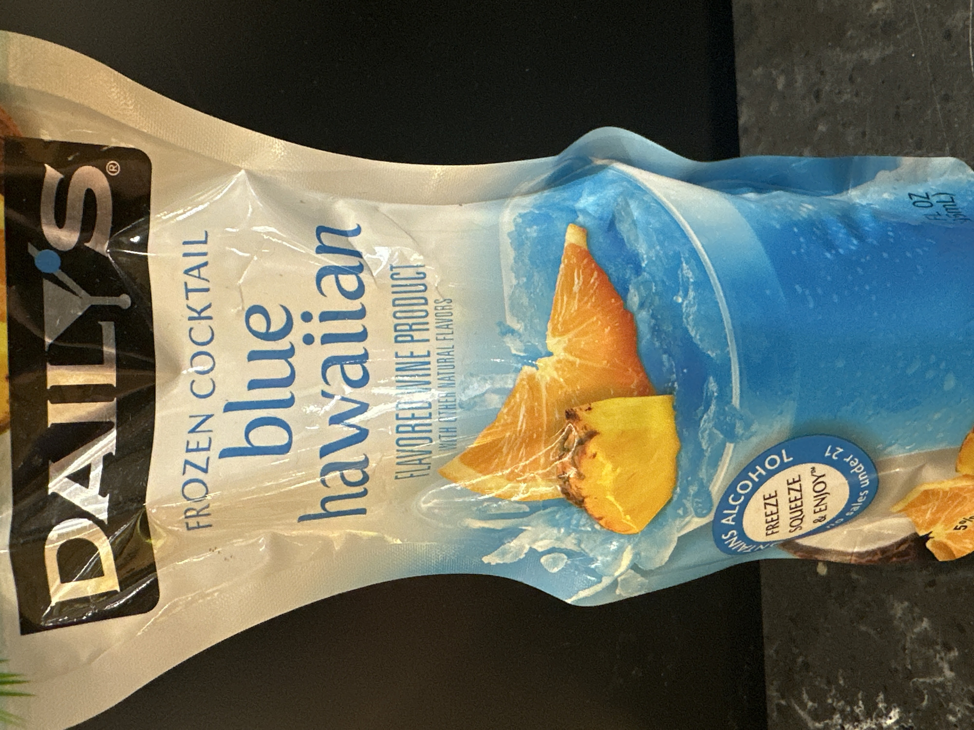 A pouch of "DAILY'S FROZEN COCKTAIL blue hawaiian" is photographed. The pouch is mostly white with blue graphics depicting a frozen beverage and fruit slices of orange and pineapple. Text on the pouch includes "DAILY'S", "FROZEN COCKTAIL", "blue hawaiian", "FLAVORED WINE PRODUCT", and "WITH OTHER NATURAL FLAVORS". A circular blue label on the lower left side of the pouch contains text that reads "TAINS ALCOHOL", "FREEZE SQUEEZE & ENJOY™", and "No sales under 21". A portion of a coconut is visible behind the circular label. The pouch is resting on a dark countertop. The background is a dark, out-of-focus wall. The image is taken in Camp Hill, United States of America.