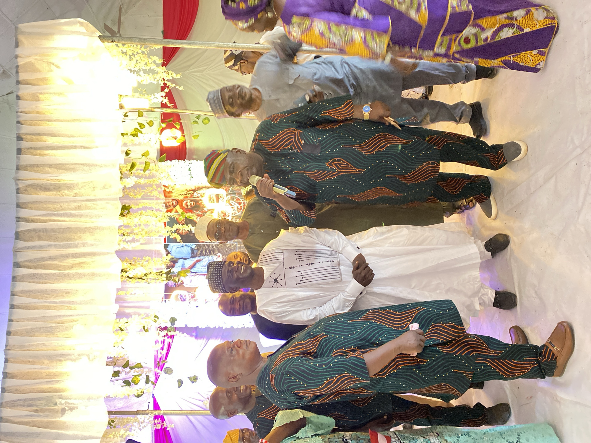 Here is a description of the image:
The image appears to be taken at an event, possibly a wedding or celebration, in Dutsen Kato, Nigeria. It shows a group of men standing in a line, seemingly addressing the audience or posing for a photograph. The men are dressed in traditional Nigerian attire, including colorful robes and hats. One man is holding a microphone. The background features decorations, including white and red drapery, and artificial flowers. The floor is covered in a white material, possibly fabric or a carpet. The setting indicates a festive occasion.