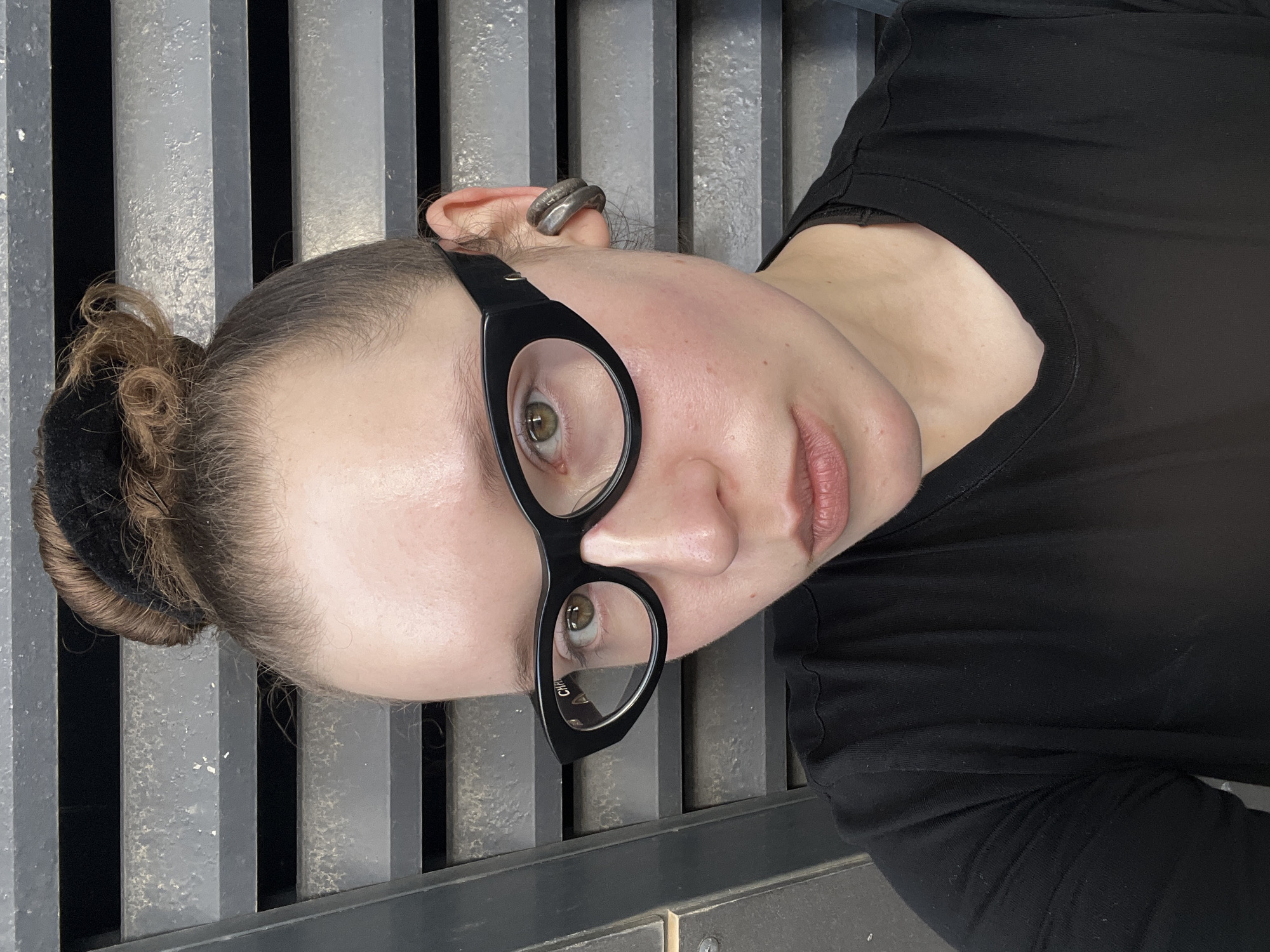 A person lies supine on a grey metal grate. The person has light skin, green eyes, and dark hair pulled into a bun. They wear black-rimmed glasses and a black shirt. A ring is visible in the person's right ear. The location is presumed to be Tel-Aviv, Israel.