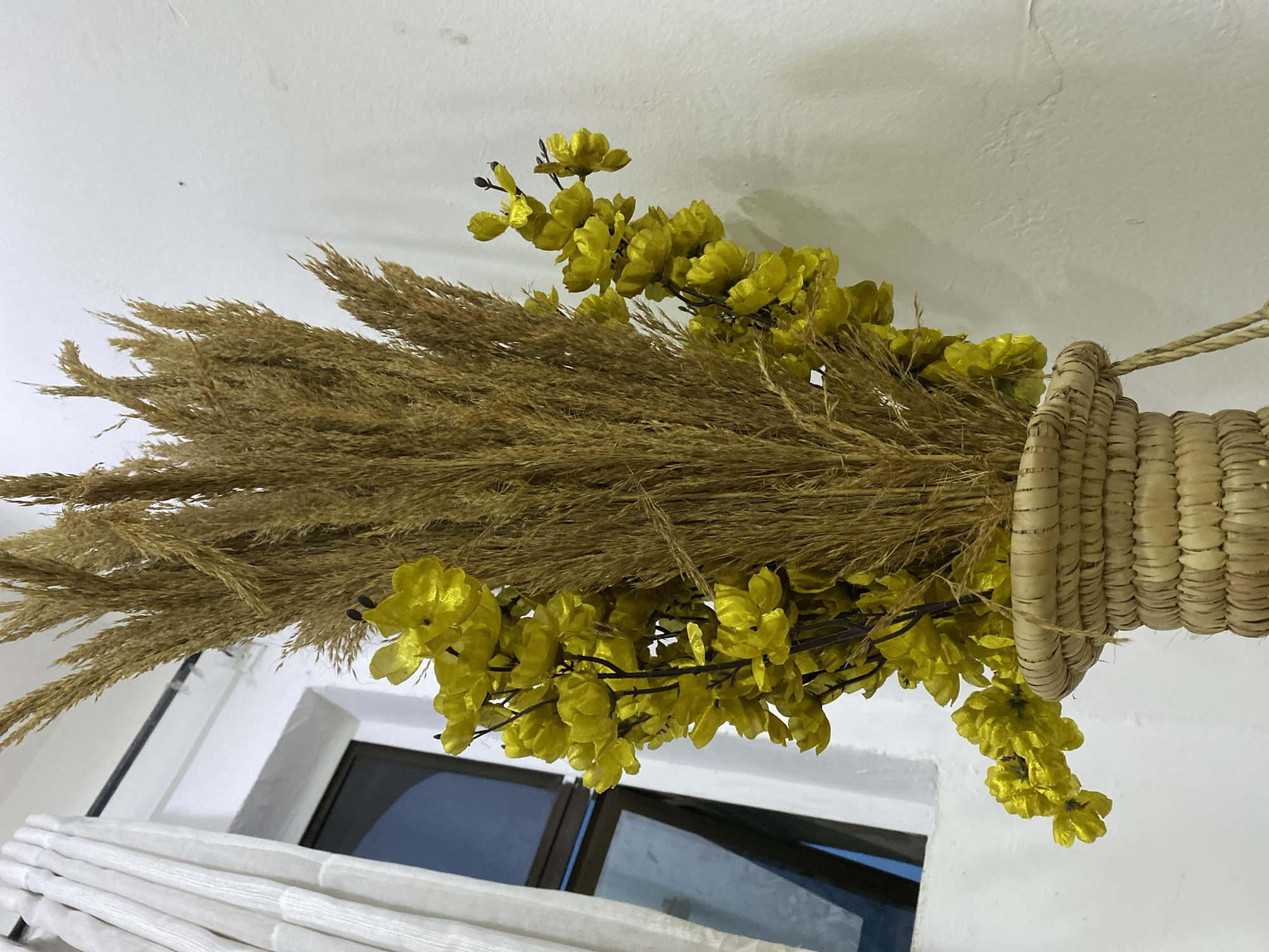 This media file captures an indoor scene featuring a decorative arrangement suspended from a white ceiling. The primary subject is a large bundle of light brown, feathery dried grass, possibly pampas grass, intricately combined with sprigs of artificial bright yellow flowers. This botanical display is held together by a thick, cylindrical woven element, resembling a light-colored straw basket or a tightly bound rope, from which a thin string hangs, indicating its attachment point to the ceiling.
Below this overhead decoration, a window is partially visible in the lower half of the frame. It features white frames and dark glass, suggesting it might be reflecting the indoor environment or looking out onto a shadowed area. In the bottom left corner, a portion of a white, horizontally pleated curtain or blind is also visible. The walls and ceiling are painted a clean white, providing a bright and neutral backdrop for the natural-toned decoration.
There are no people present, and no specific activity or event is taking place; the image presents a static interior view. The lighting appears soft and diffused, possibly indicating an indoor light source or an overcast day outside. No visible text is present in the image. While the location context is given as Badagry, Nigeria, there are no explicit visual cues within the image itself to confirm this geographical location. The aesthetic suggests a rustic or natural decorative style within a modern or traditional setting.