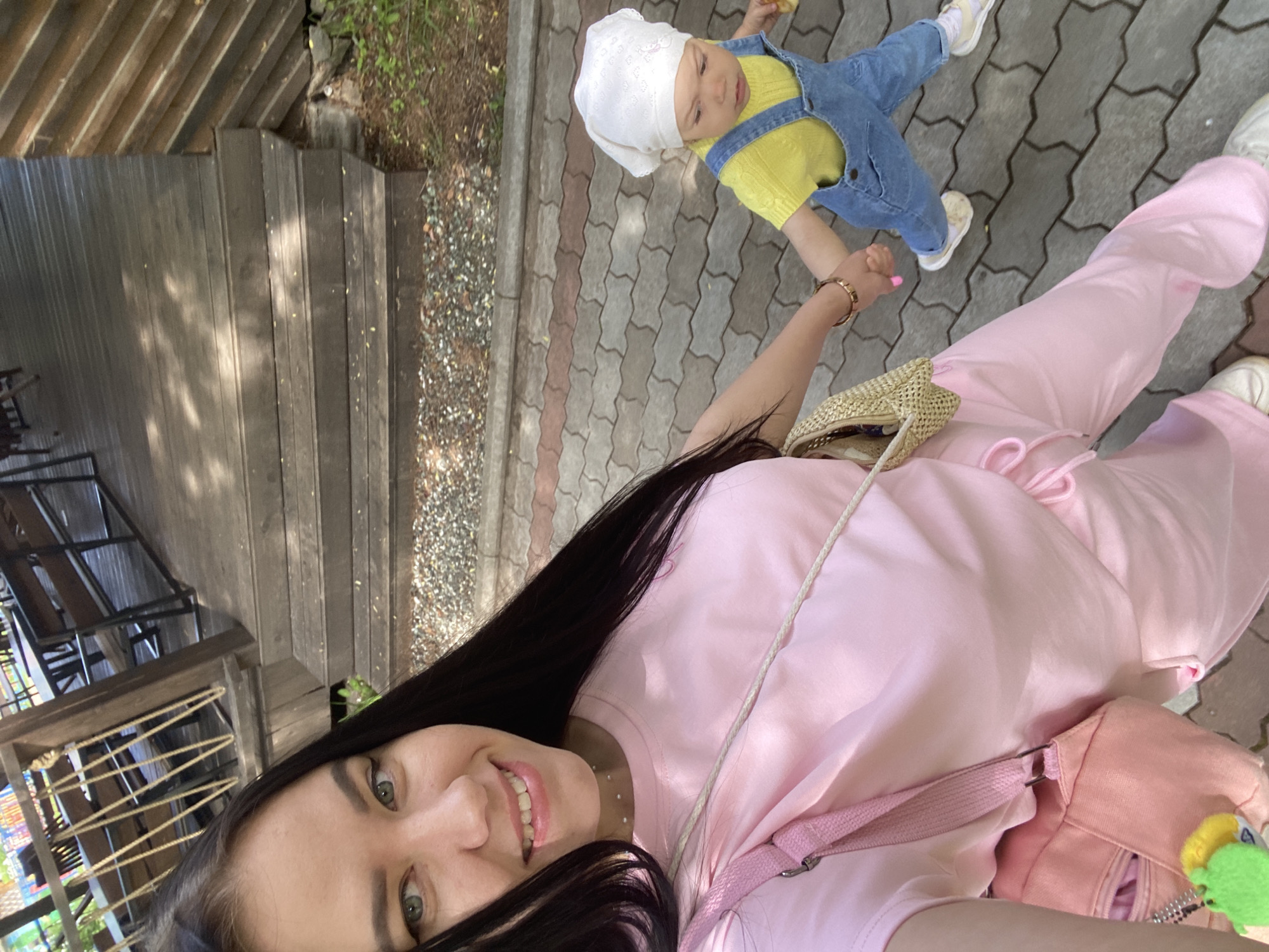 An outdoor, rotated selfie captures a woman and a small child holding hands. The woman, with long dark hair and green eyes, smiles broadly at the camera. She is wearing a light pink tracksuit and has a small, light brown woven bag with a colorful green and yellow toy charm attached. On her wrist, she wears a gold bracelet. The child, looking slightly upwards and to the right, wears a white bonnet, a bright yellow top, and denim overalls.
They are positioned on a paved pathway composed of interlocking grey and reddish-brown pavers. To their left, there is a dark wooden structure, possibly steps or decking, with some gravel and sparse greenery visible next to it. The background on the far left shows more of the wooden structure and what appears to be part of an outdoor seating area. The image suggests a bright, sunny daytime setting, with no indication of bad weather. No text is visible in the media. The scene depicts a casual outing in Chaikovsky, Russia.