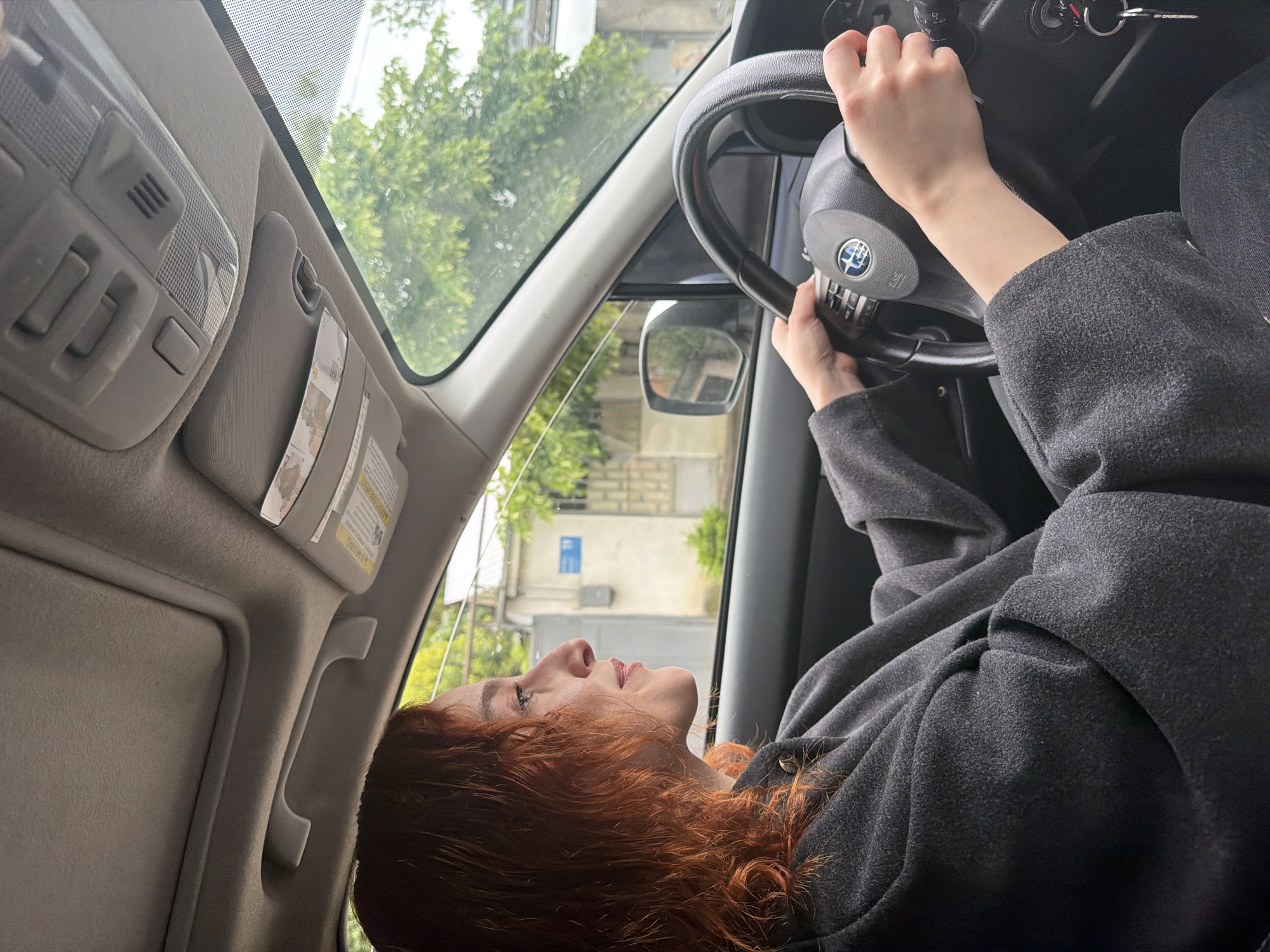An upside-down, angled shot reveals a fair-skinned woman with red hair at the wheel of a vehicle, likely a Subaru, as indicated by the steering wheel logo. She is wearing a dark gray coat and has her hands on the steering wheel. Outside the car window, a building is visible. Based on the street sign number 10 on the building, it's possible the location is Tbilisi, Georgia. The interior of the car includes the roof console and a portion of the sun visor. The keys are in the ignition.