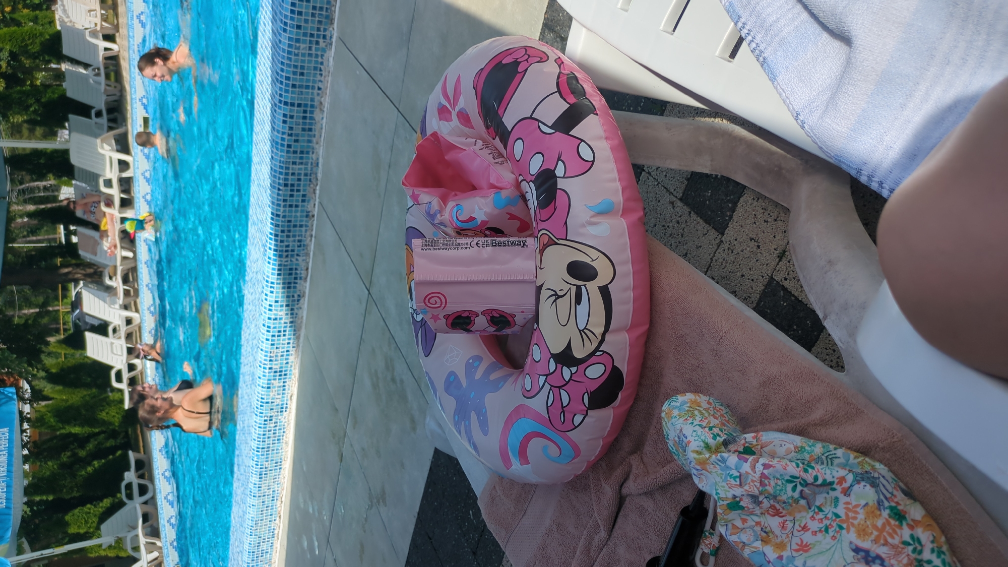An outdoor swimming pool area in Vadul lui Voda, Moldova, is depicted. In the foreground, a pink inflatable swimming ring featuring Minnie Mouse characters rests on a light pink towel, which is laid out on a white lounge chair. A baby's sun hat with a floral pattern is placed next to the ring on the towel. Beyond the lounge chair, a swimming pool with blue water is visible. Several people are in the pool, including two women in black swimsuits, one with braided hair, and a child wearing a green floatation device. In the background, white lounge chairs are arranged, and lush green trees and foliage surround the pool area. The ground surrounding the pool is paved with grey and black square tiles.