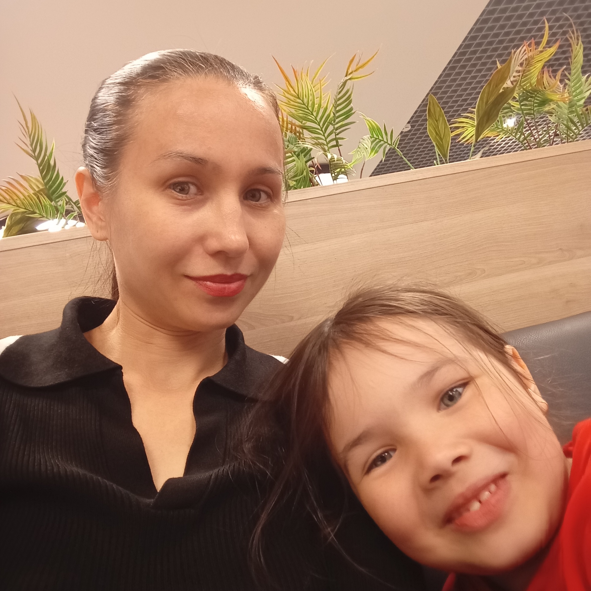 This is a close-up, indoor selfie of two individuals, likely a mother and daughter. The woman on the left is an adult with her hair pulled back, wearing a black collared shirt. She has light skin and is looking towards the camera with a slight smile. The child on the right, resting their head on the woman's shoulder, has fair skin and brown hair, with their eyes wide and a joyful expression. They are wearing a red garment. The background consists of a light wood-paneled wall and decorative artificial plants on a shelf above. The lighting appears to be artificial. There is no identifiable information about the specific location within the city of Izhevsk, Russia.