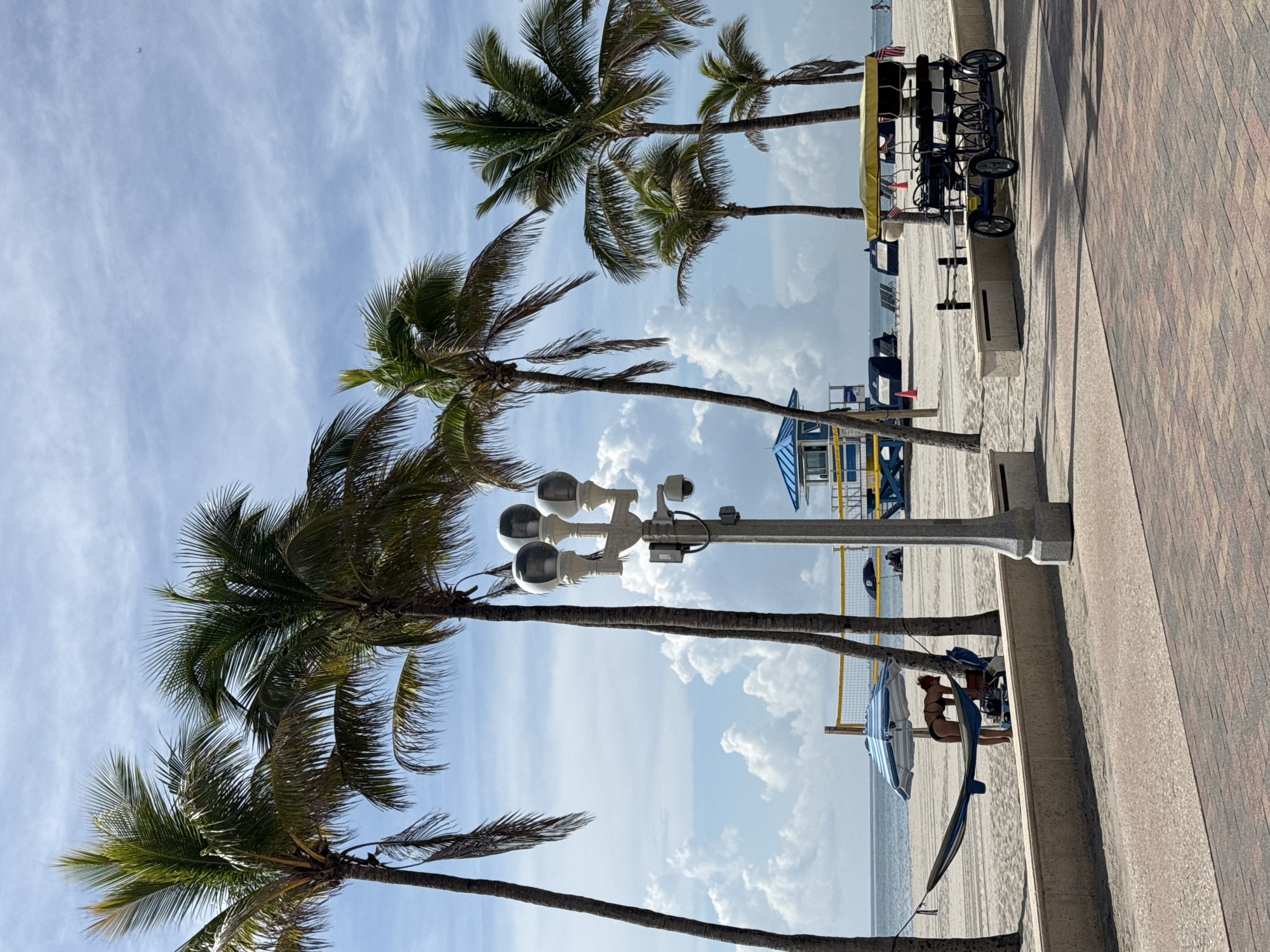 The image depicts a sunny day at the beach, likely in Hollywood, Florida, given the presence of palm trees and the characteristic beach town atmosphere. The scene is a boardwalk overlooking a sandy beach with the ocean in the background. Several palm trees are prominent in the foreground, their fronds swaying gently against a bright blue sky dotted with fluffy white clouds.
On the boardwalk, a multi-person pedal cart with a yellow canopy is parked. It appears to be a recreational vehicle for rent, a common sight in tourist beach areas. Further down the boardwalk, a lamp post with multiple lights and security cameras stands tall.
On the beach, a volleyball net is set up, suggesting recreational activities. Beneath a striped umbrella, a person in a bikini is visible, appearing to be stretching or performing yoga. Another person is seated nearby, and a surfboard is propped up. The overall mood is relaxed and leisurely, indicative of a vacation or a pleasant day outdoors. The time of day appears to be morning or afternoon, with ample sunlight casting shadows and highlighting the vibrant colors of the scene. The paving stones of the boardwalk are in the foreground, adding a textural element to the image.