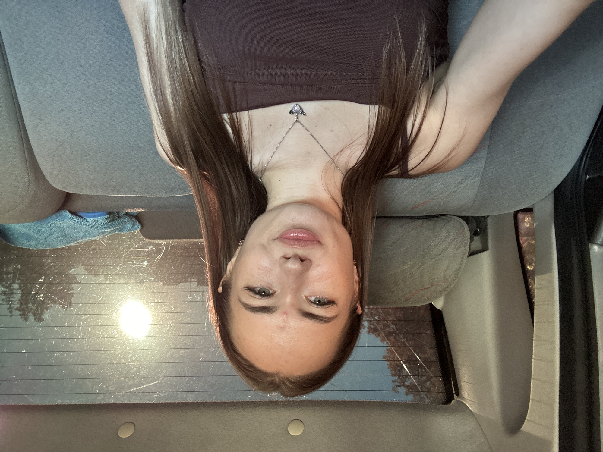 A young Caucasian woman with long brown hair sits in the back seat of a car. She is wearing a brown strapless top and a silver necklace with a winged heart pendant. The sun is shining through the rear windshield, creating a bright glare and casting a warm light on the scene. The car's interior is visible, with gray fabric seats and a textured headliner. Outside the car, trees and what appears to be a cityscape are visible through the dirty windshield. The woman is looking directly at the camera with a slight smile.