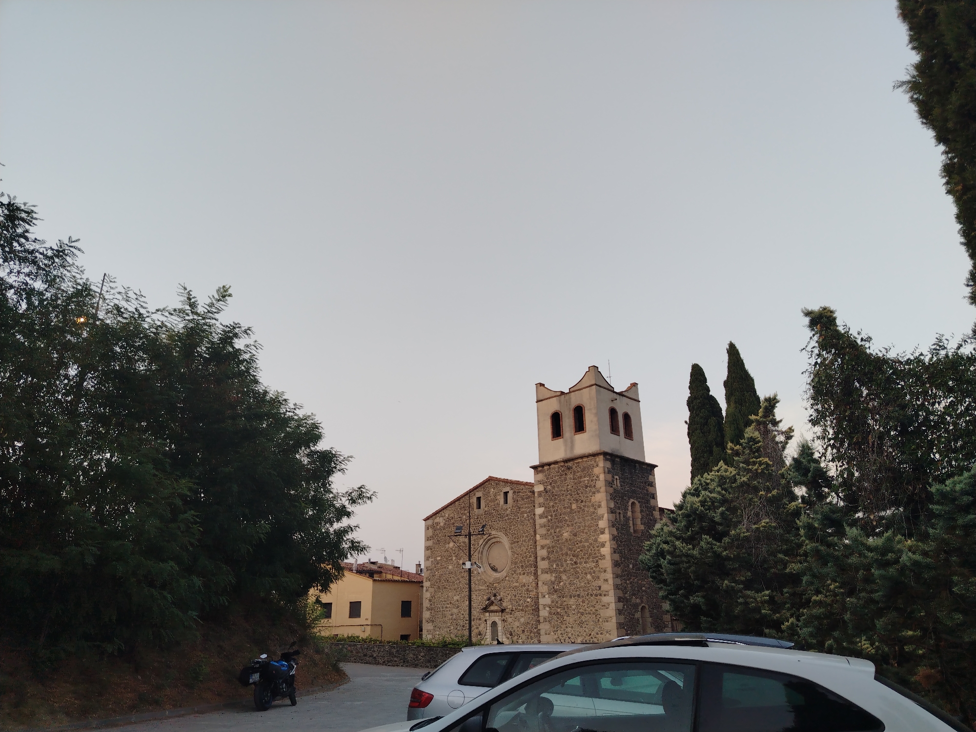 This media file depicts a serene scene in Hostalric, Spain, likely during the late afternoon or early evening as suggested by the soft, diffused light and the pale sky. The focal point is a stone church with a distinct bell tower, characterized by its rustic stone construction and arched openings at the belfry. Adjacent to the church is a building with a light yellow facade and tiled roof, hinting at a residential or community area.
In the foreground, two cars are parked, one silver and the other a lighter color, partially obscuring the view of the church. To the left of the frame, a winding road leads uphill, bordered by lush green trees. A motorcycle with luggage strapped to it is parked on the side of this road, suggesting recent travel or a stop by a visitor. Several tall, conical trees, possibly cypresses, stand to the right of the church, adding to the Mediterranean feel of the location.
The overall atmosphere is peaceful and quiet, with no visible people, implying a moment of calm in the town. The weather appears clear and mild. The stone textures of the church and the surrounding greenery are prominent details, contributing to the picturesque setting. No visible text is present in the image.