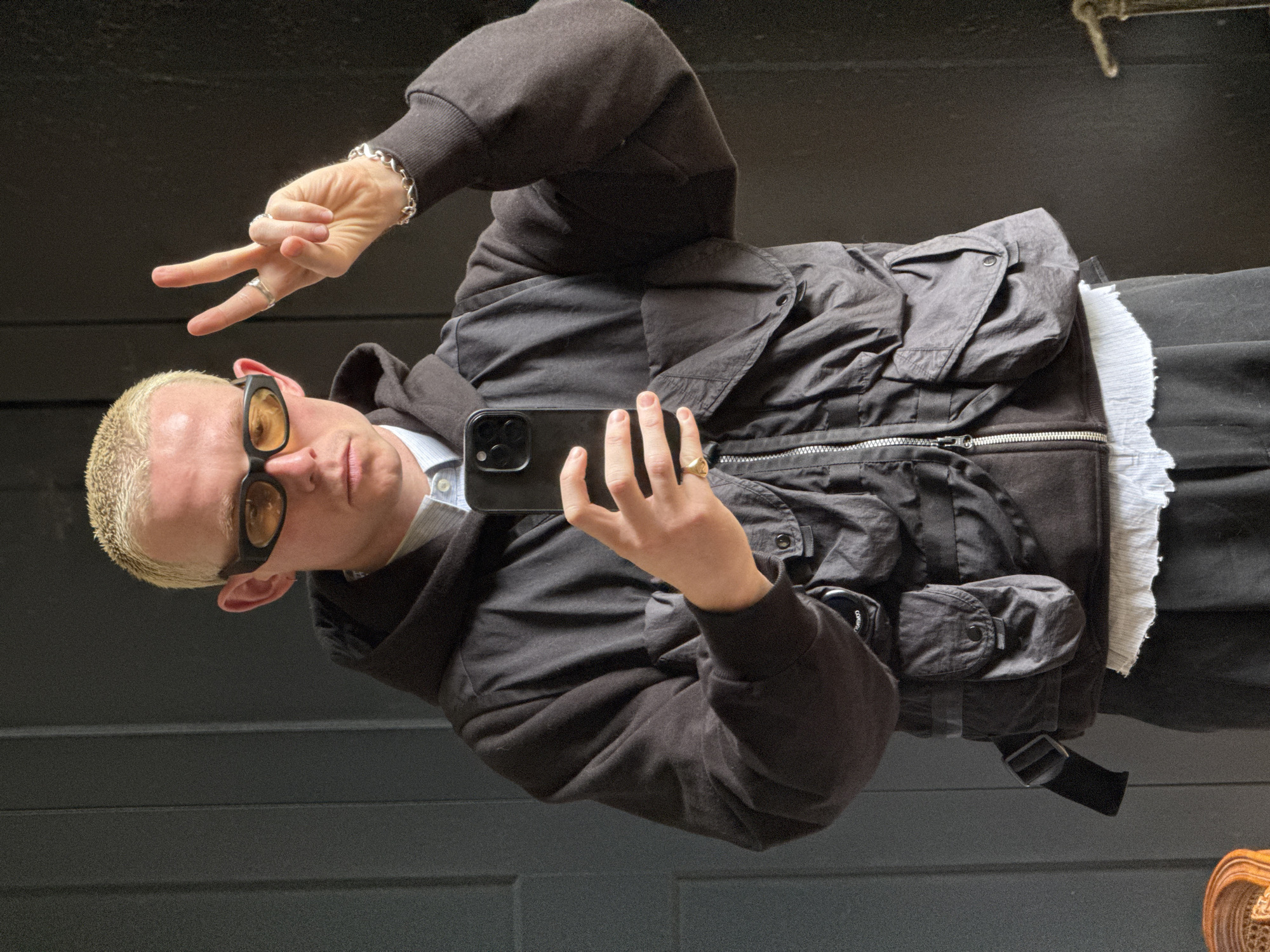 The image, rotated 90 degrees clockwise, captures a person taking a mirror selfie. The subject is a young male with short, bleached blond hair and light skin. He is wearing yellow/orange tinted sunglasses with thick black frames, a silver chain bracelet on his right wrist (viewer's left), a silver ring on his right index finger, and a gold-toned signet ring on his right ring finger. He holds a black smartphone (likely an iPhone due to the camera array) in his right hand, angled towards himself, while his left hand is raised, forming a peace sign.
He is dressed in a dark grey or black hooded sweatshirt over a light blue collared shirt. Over the sweatshirt, he wears a dark grey or black technical jacket, possibly made of nylon or a similar synthetic material. The jacket features multiple pockets, including a distinct circular patch with a lens on the left sleeve pocket, characteristic of C.P. Company. It also has a prominent silver zipper down the front. A white, textured garment or lining is visible beneath the jacket on his right side, and dark trousers complete the outfit.
The background is a plain, dark surface, likely an indoor wall. No specific location cues for London are visible, other than the provided context. The person's expression is neutral, and the gesture conveys a casual or friendly mood. The lighting suggests an indoor setting, and the time of day or weather cannot be determined.