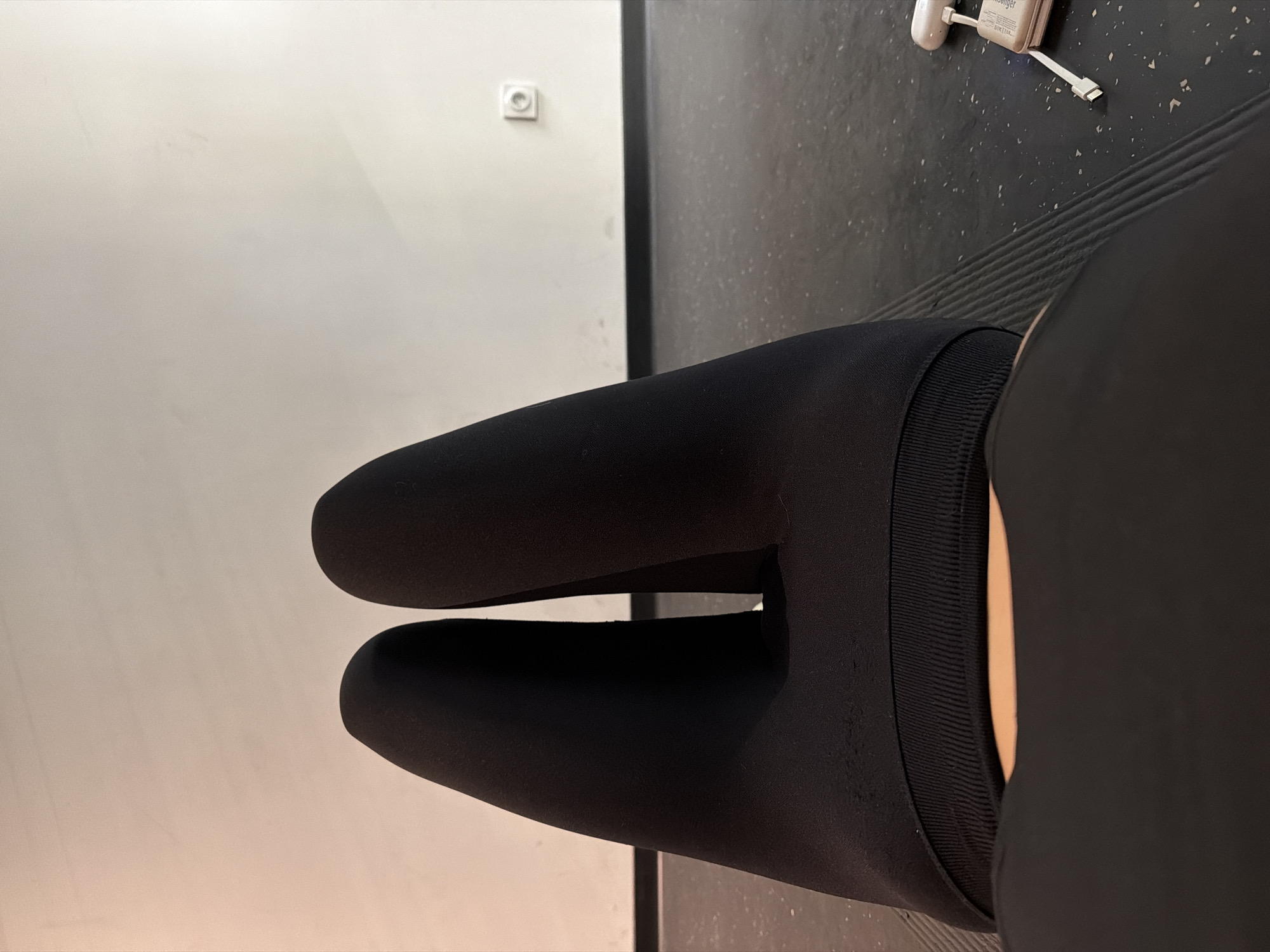 The image presents a close-up, high-angle view focusing on a person's thighs and knees clad in black leggings. The texture and contours of the fabric are emphasized, revealing the shape of the legs beneath. The person is situated indoors, with a segment of a charger visible in the background, suggesting a gym environment. The background includes a light-colored wall and the edge of a dark floor. The image lacks any distinct identifiers, so Paris cannot be definitively determined.