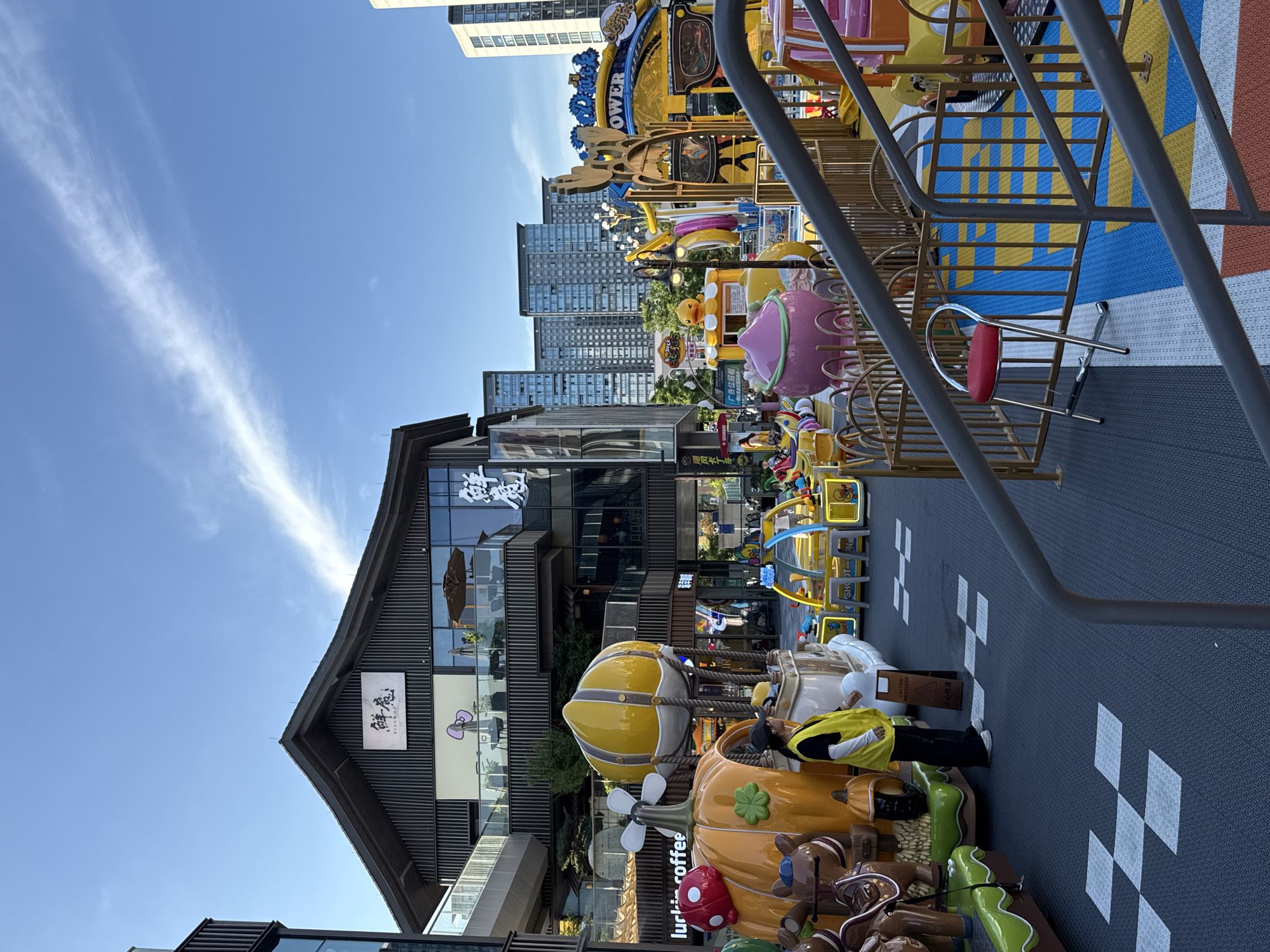 This is a photograph taken in Yiwu, China, depicting an outdoor amusement area, possibly a theme park or entertainment zone, adjacent to commercial buildings. The scene is set under a clear blue sky with a wispy cloud formation. In the foreground, a grey metal railing and stairs lead into the amusement area.
The amusement area is filled with colorful rides and attractions. Prominent among these are several whimsical, cartoonish structures. One ride features a large, yellow hot air balloon-like sphere. Nearby, a large orange pumpkin-shaped structure with attached cartoon animal figures (bears) is visible, along with a red mushroom. Several other smaller rides and play structures with bright colors like yellow, pink, and blue are scattered throughout the area. A small red stool is placed near a golden-colored fence. The ground in the amusement area has a checkered pattern of white squares on a dark blue textured surface, interspersed with sections of yellow, blue, and orange paving.
Behind the amusement area, a multi-story building with a dark, traditional-style roofline stands out. This building has large windows and balconies, and signage with Chinese characters, one of which appears to be "鲜鼎" (Xiān Dǐng). Other commercial establishments are visible, including a sign that reads "luckin coffee". Further in the background, several modern high-rise residential or office buildings are visible, indicating an urban setting.
Two individuals are present in the scene. One person, wearing a yellow vest over dark clothing and a cap, is standing near the pumpkin ride. Another person is partially visible walking in the background near a building entrance. The overall impression is a lively and family-friendly outdoor entertainment space integrated within a modern urban commercial district.