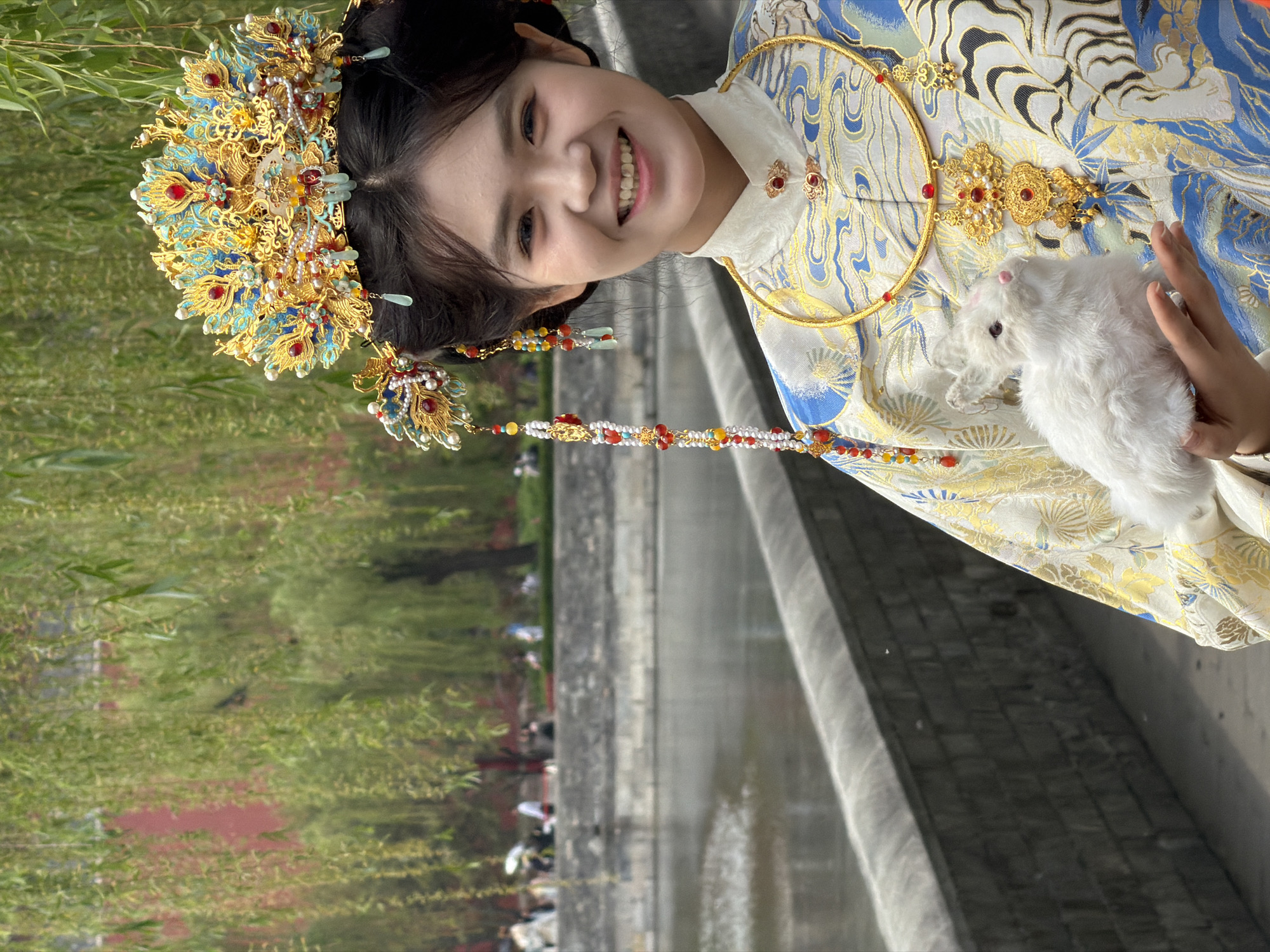 A medium shot depicts a woman, smiling directly at the camera, holding a small, white rabbit in her arms. She is adorned in an elaborate, traditional Chinese headdress of gold and blue, with cascading beaded strands. Her clothing is also traditional, featuring a high collar, and a pattern of swirling gold and blue designs on a white background. A gold neck ring and ornamental clasps complete her ensemble.
The backdrop suggests an outdoor setting. A gray stone wall runs along the bottom right of the frame, with a body of water visible behind it. In the distance, trees with green foliage are seen, along with some people. The scene appears to be taken in Dongcheng District, China.