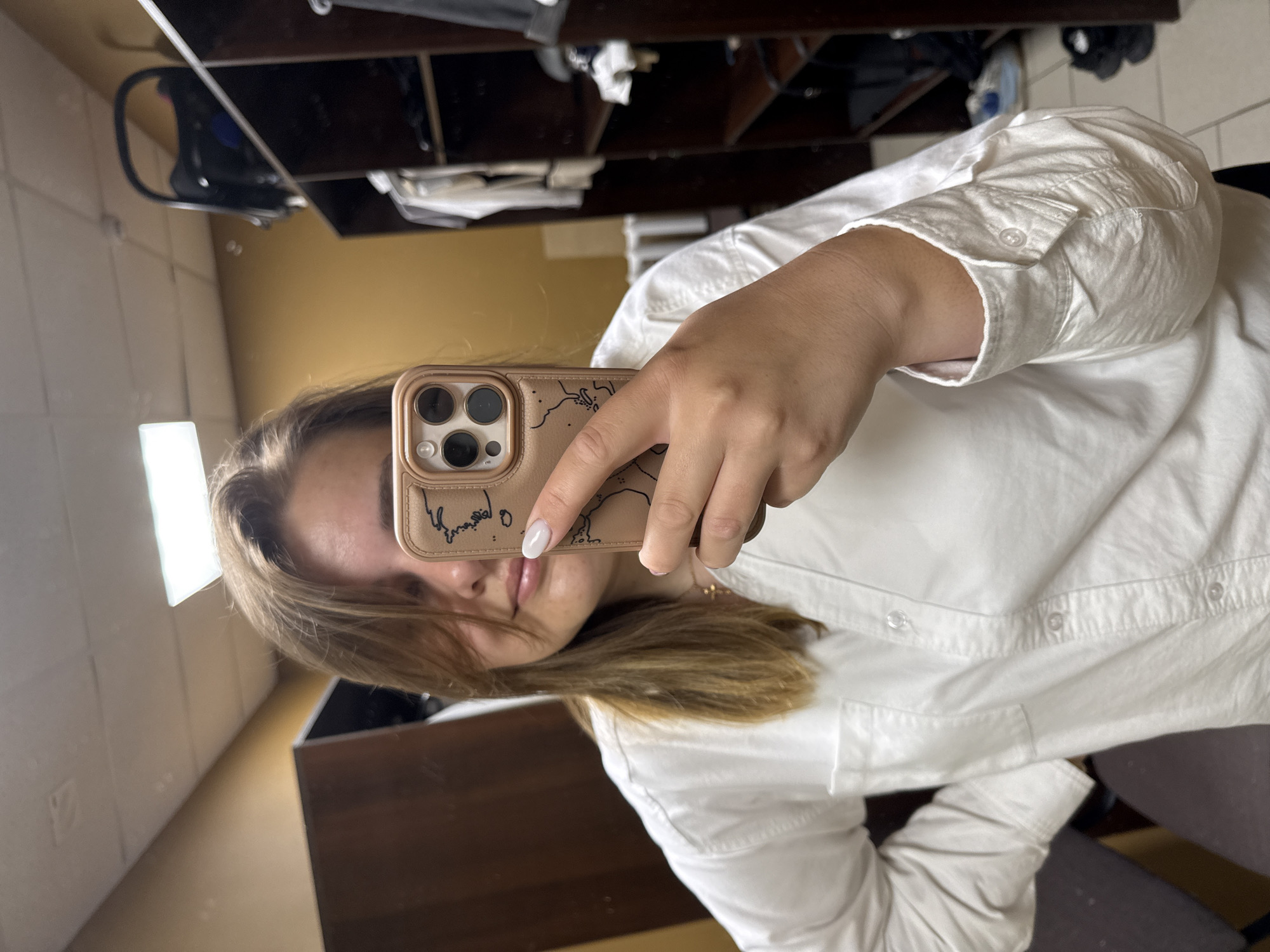 A young woman is taking a selfie in what appears to be a bathroom. She is wearing a white button-down shirt and holding a phone with a tan-colored case. The background includes a mirror reflecting a shelf with various items. The overall environment suggests an interior setting. There is no evidence to link this picture to the city of Chaikovsky, Russia.