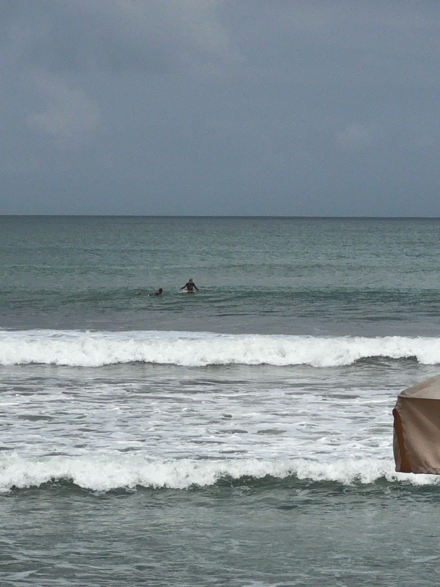 The image depicts a scene at Kuta Beach, Indonesia, under a cloudy sky. The ocean occupies the majority of the frame, with several waves breaking closer to the shore. In the mid-distance, two individuals are visible in the water. One person is sitting on a surfboard, while another appears to be swimming. In the foreground, a portion of a beach umbrella is visible on the right side of the image, suggesting the presence of a beach setup. The overall atmosphere is subdued, likely due to the overcast weather.