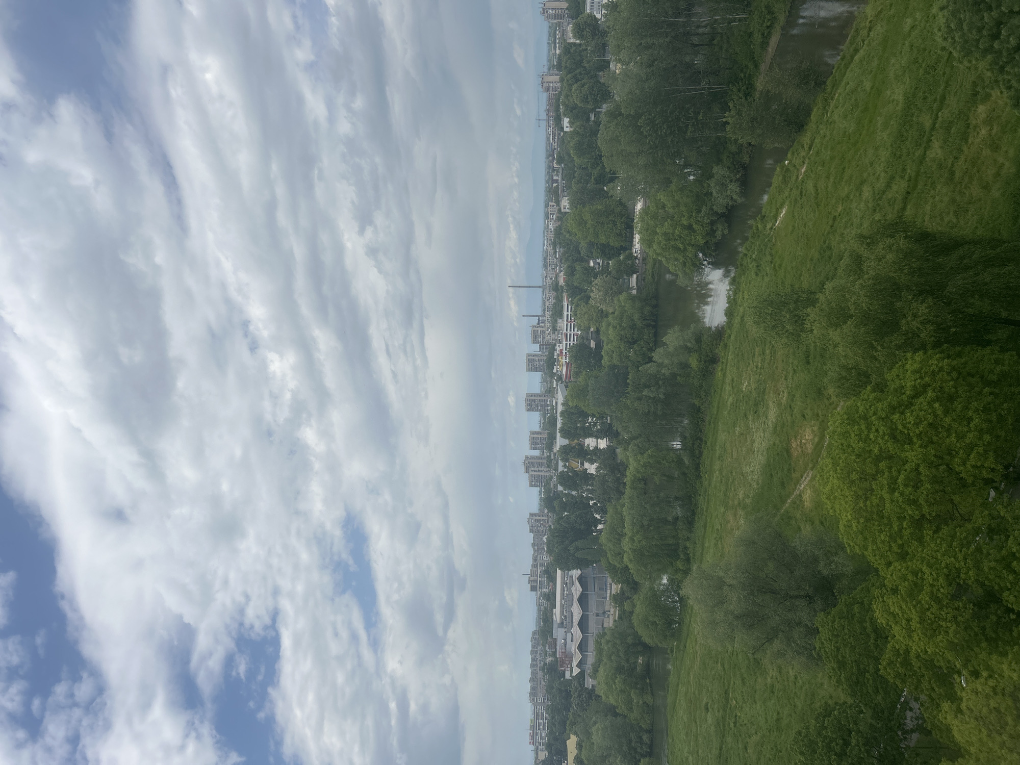 An aerial view shows a city landscape under a sky filled with white and gray clouds. Buildings are visible along the horizon. A river and a green, grassy embankment run alongside the urban area. Trees dot the scene.