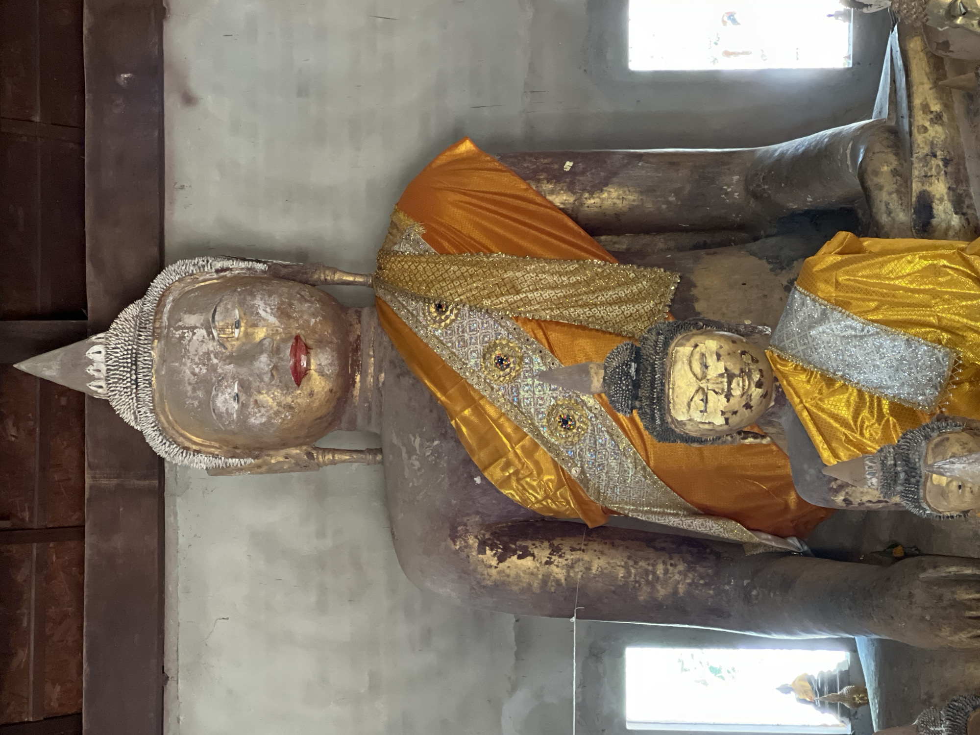 This image shows two large Buddha statues inside a building. The statues are adorned with yellow and silver robes. The larger statue is behind a smaller one, and both appear to be made of stone and have gold leaf accents. The environment is dimly lit with natural light coming from windows on either side of the statues. The walls are a rough, textured gray. The roof appears to be made of dark wood. The location is identified as Baan Harn, Thailand.