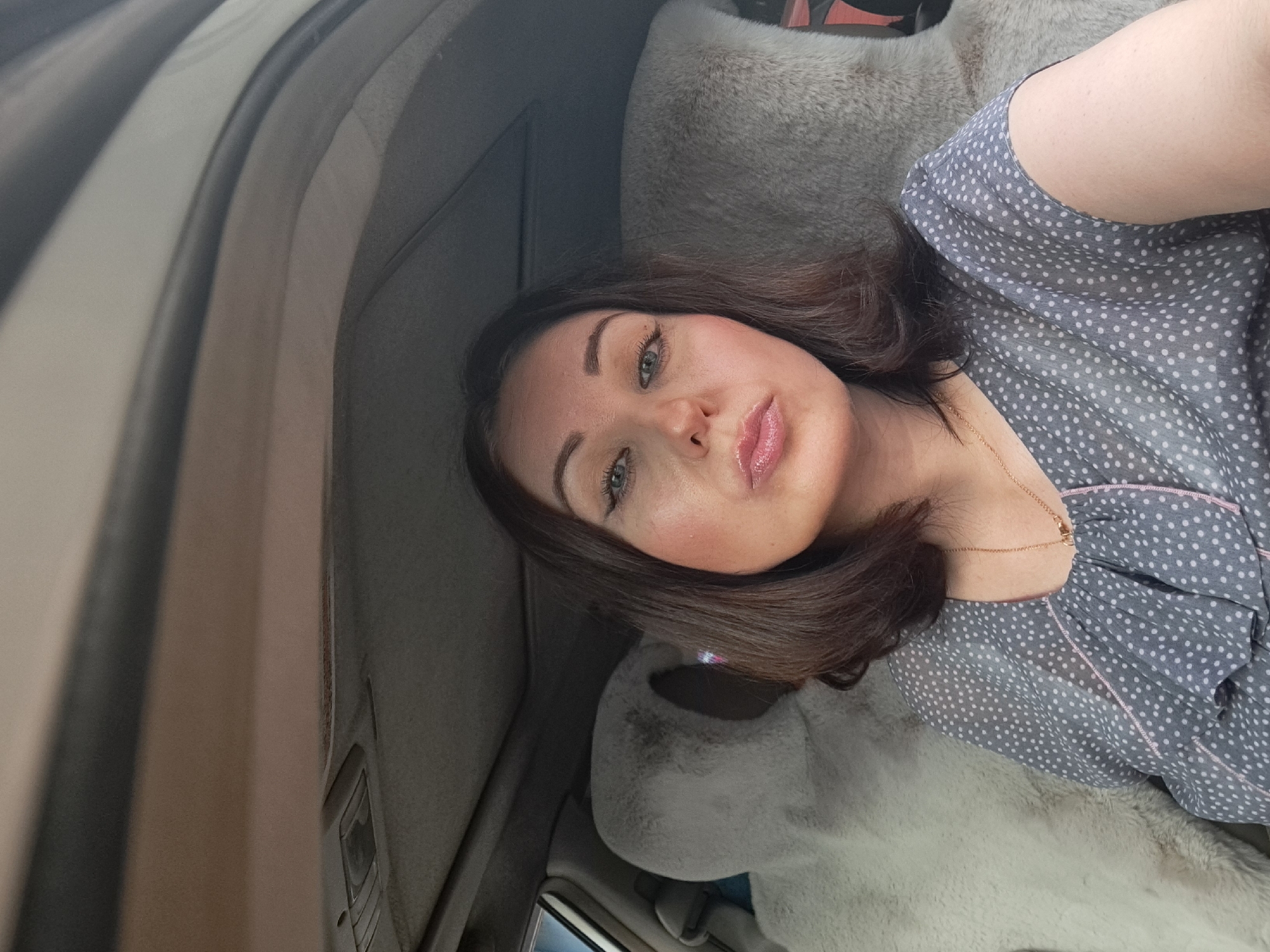 A selfie, taken from a low angle inside a car, shows a woman with brown hair and fair skin. She is wearing a gray polka-dot blouse with pink trim and a gold necklace. Her makeup includes winged eyeliner and light pink lipstick. Her head is tilted slightly, and her lips are pursed. Behind her, the plush, light-colored headrests of car seats are visible. The car's sunroof is open, revealing a gray sky. The scene is indoors, within the confines of a vehicle.
