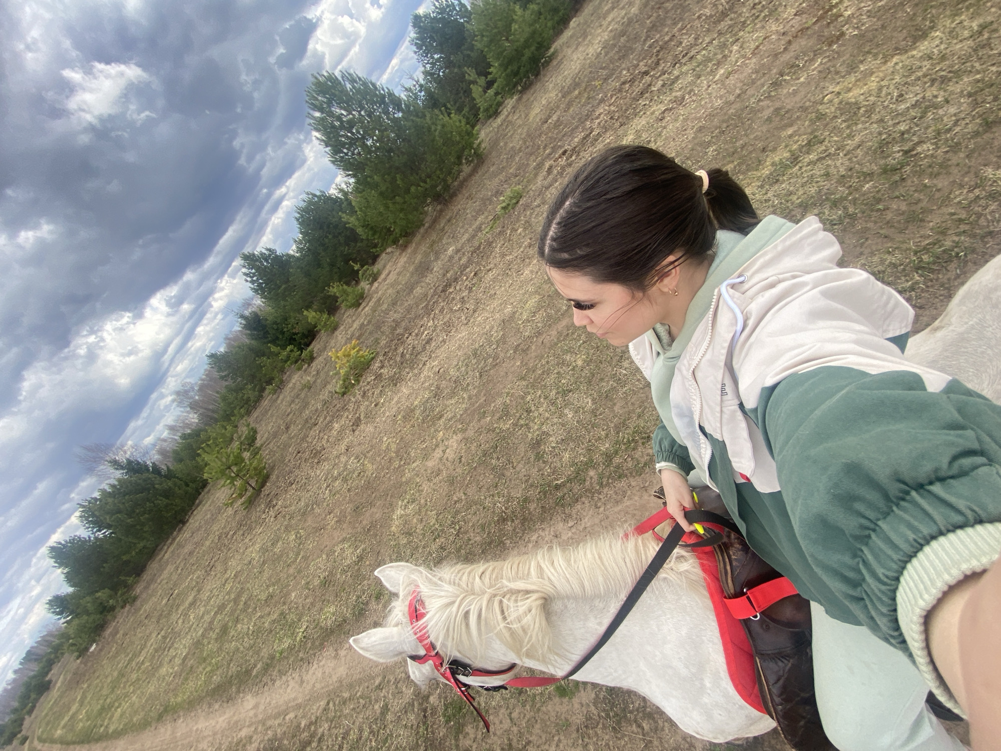 A woman is riding a white horse in a field. The field is dry and grassy. There are trees in the background. The sky is cloudy. The location is Zhigalki, Russia.