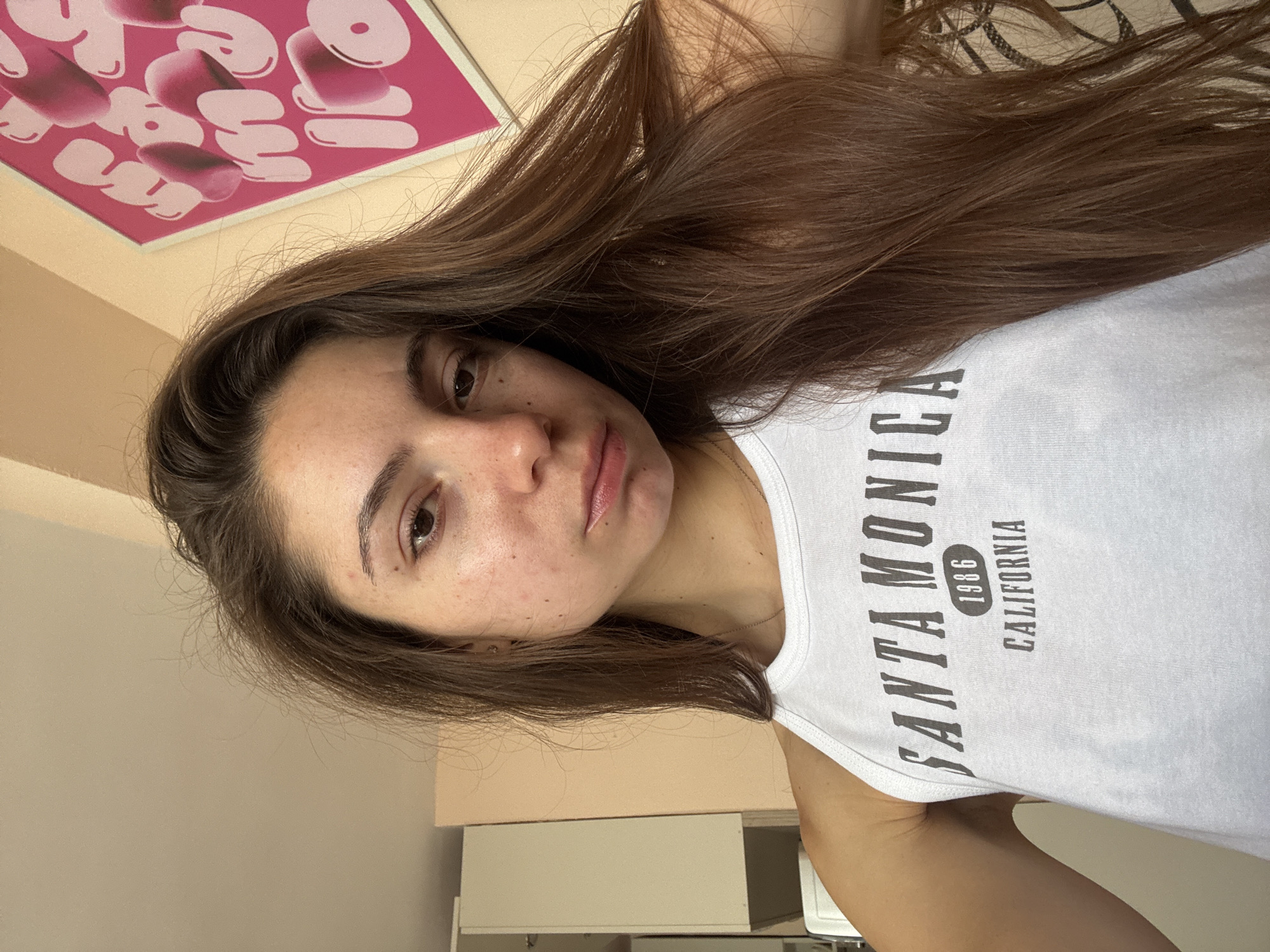 A young woman with long, brown hair is taking a selfie in what appears to be a room. She is wearing a white tank top with "SANTA MONICA 1986 CALIFORNIA" printed on it. The background shows a wall with a framed pink artwork and some furniture, possibly a cabinet. The lighting suggests it might be daytime. The woman's facial expression is neutral, and she is looking directly at the camera. There is no visible indication of the city of Varna, Bulgaria, in the image.