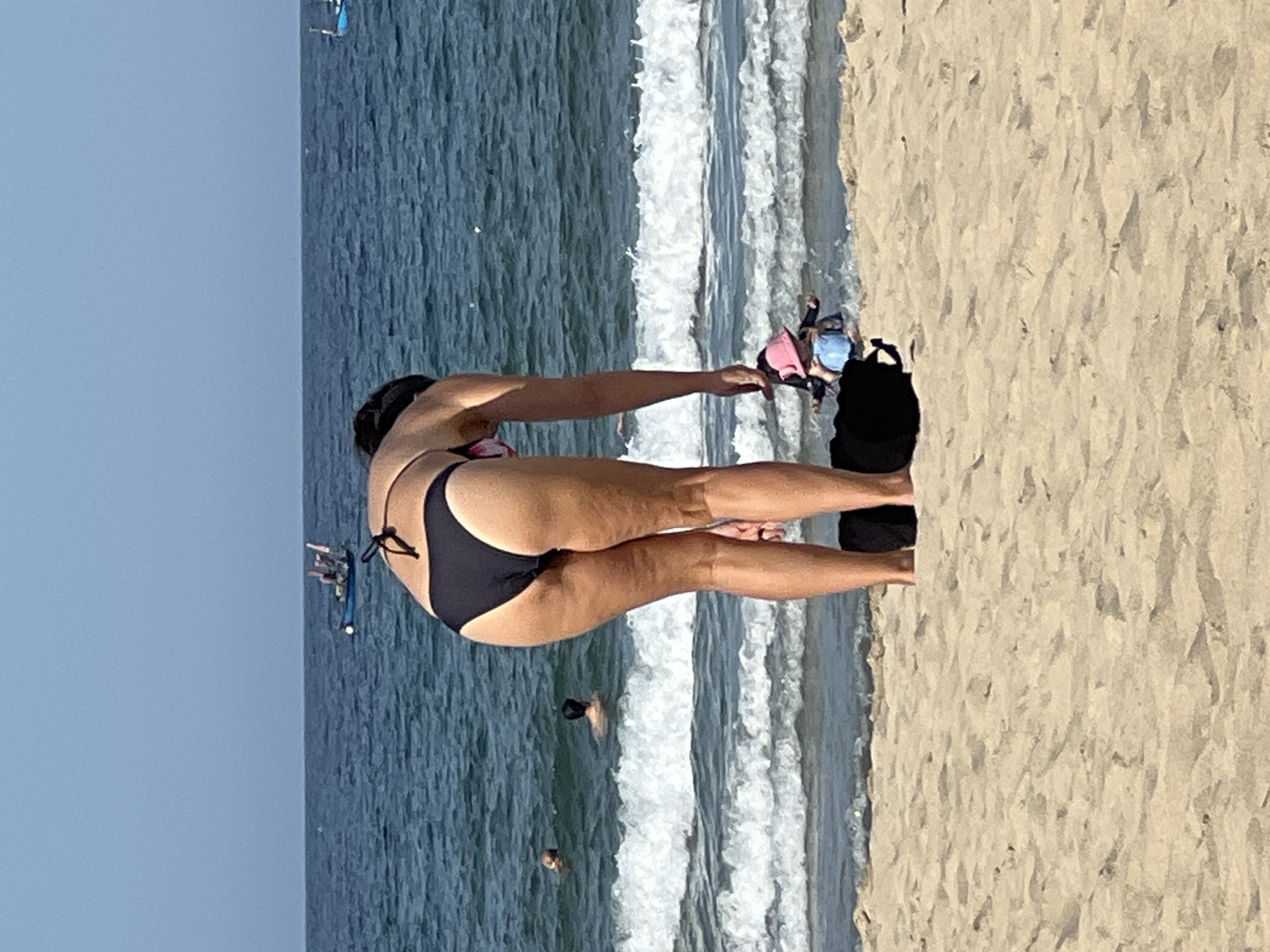 A woman wearing a black bikini bends over on a sandy beach in Đà Nẵng, Vietnam, with her back to the camera. She is facing the ocean, which is calm with gentle waves. In the distance, several small boats are visible on the water. Further out, the horizon is clear and a pale blue sky dominates the upper portion of the image. To the right of the woman, partially obscured by her, a child wearing a pink hat and a blue swimsuit is sitting on the sand, facing the water. The sand is light brown and appears dry and rippled. The overall scene depicts a typical day at the beach.