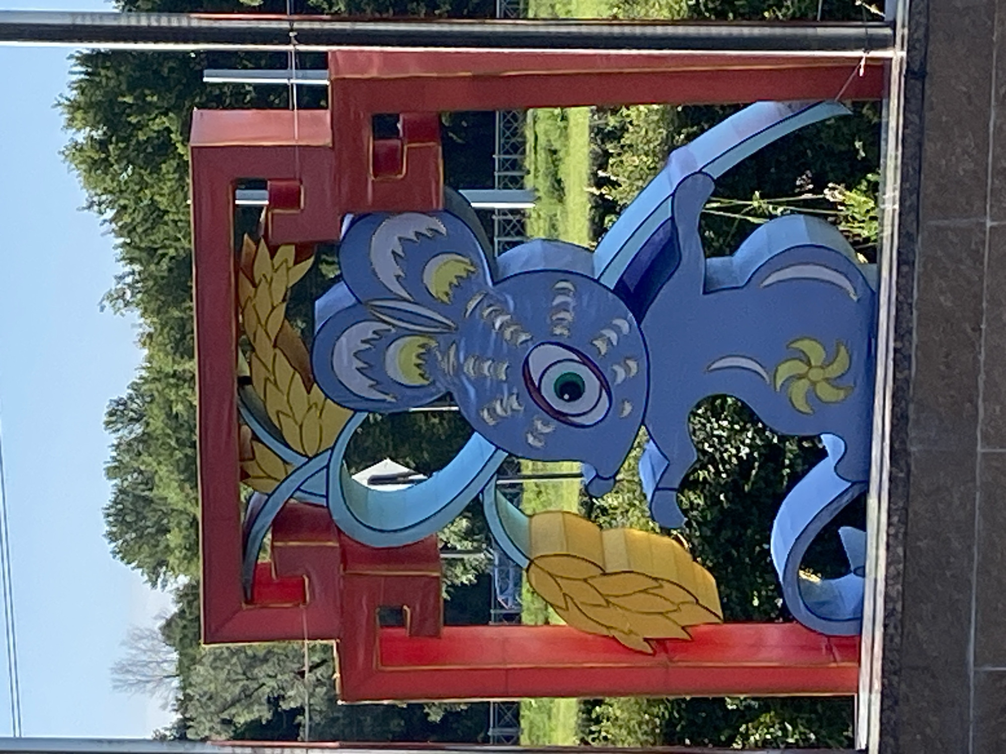 A blue mouse lantern is displayed outdoors, framed by a red, oriental-style archway. The lantern depicts the mouse holding a yellow, stylized ear of corn. The background consists of green trees and a clear blue sky. This lantern is located in the Saint Petersburg Zoo in Saint Petersburg, Russia.