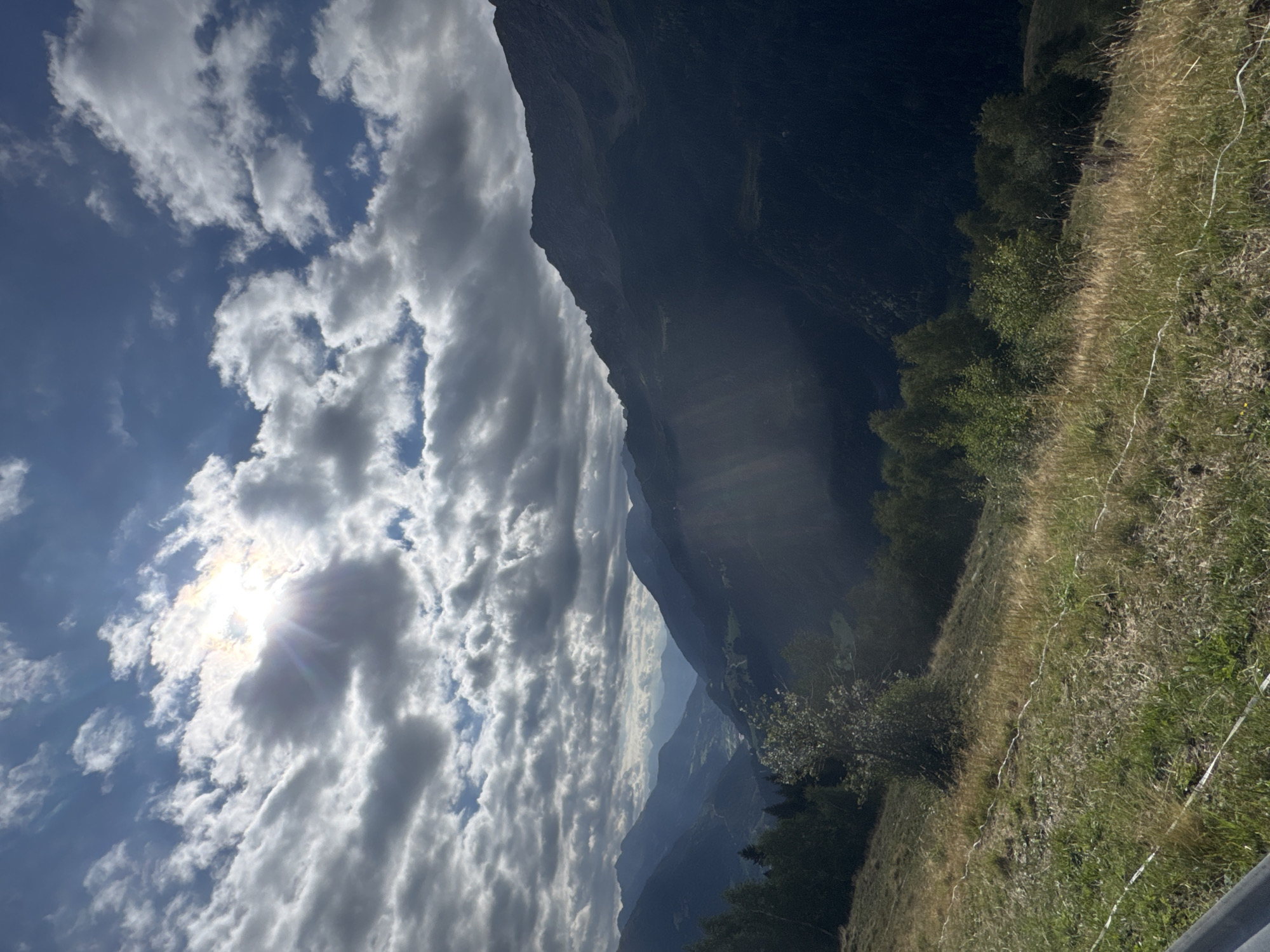 The image depicts a scenic mountain landscape, likely in the vicinity of Airolo, Switzerland. The sky is filled with dramatic, partly cloudy formations, with the sun shining brightly through a break in the clouds, creating visible sunbeams. Rolling mountains dominate the mid-ground and background, with the foreground featuring a grassy hillside. A faint white string runs horizontally across the grassy slope, possibly marking a boundary or a temporary feature. There are no people or animals visible in the frame. The overall impression is one of a natural, rugged environment under a dynamic sky.