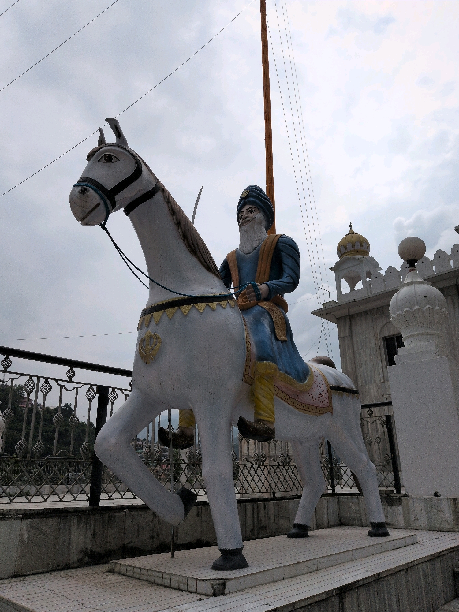 A statue of a Sikh man on horseback is shown in front of a Sikh gurdwara in Mandi, India. The statue is white with gold trim and the man is wearing a blue turban and robe. He has a long white beard and is holding the reins of the horse. The horse is white with a black mane and tail. The gurdwara is a white building with a golden dome. The sky is cloudy.