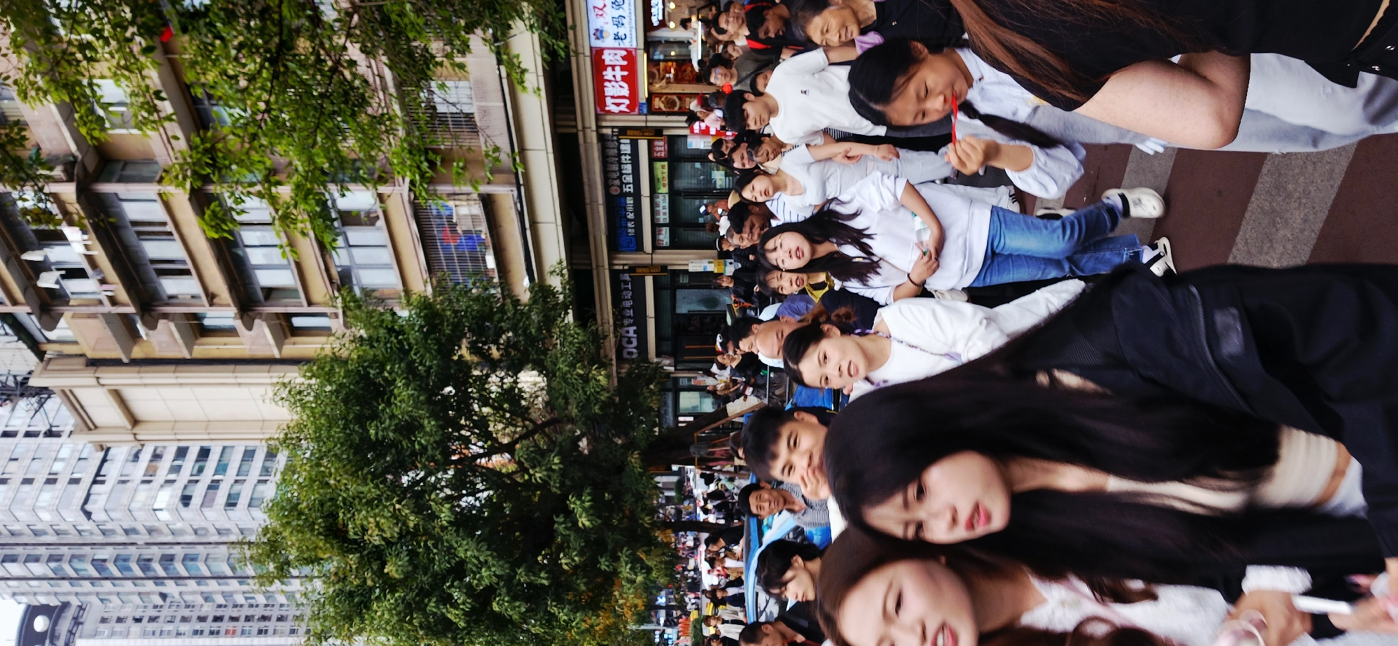 The image depicts a large crowd of people, mostly young women, gathered on a city street in Yuzhong District, China. The women are wearing similar attire, a white top and jeans. Buildings line one side of the street, with trees in the foreground. Storefront signs in Chinese are visible. Some people are interacting, while others appear to be looking at something out of the frame. The scene suggests a possible event or gathering.
