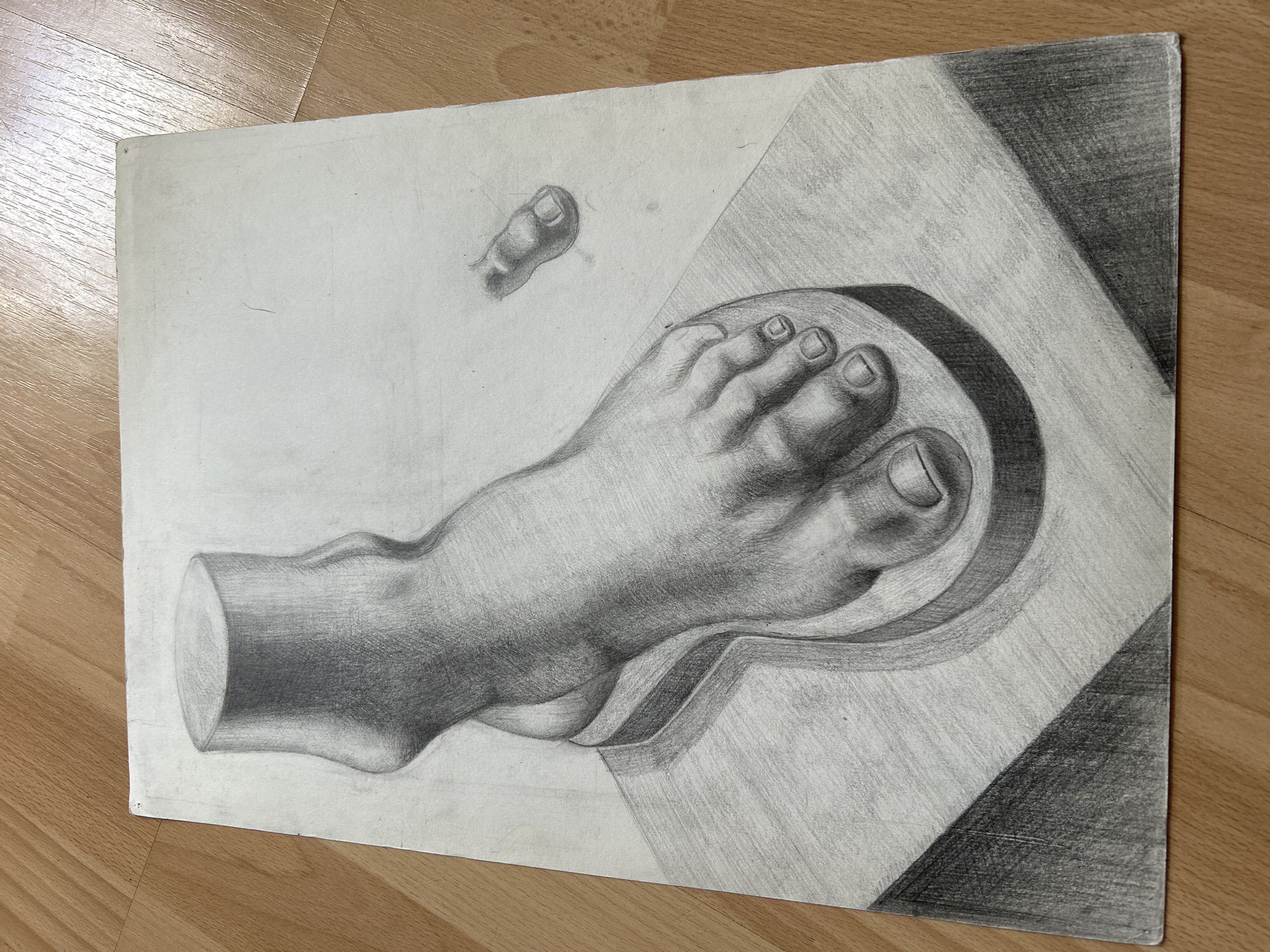 The image is a pencil drawing on paper depicting a plaster cast of a human foot and ankle. The foot is positioned within a mold, which is itself resting on a flat, rectangular surface. The drawing is rendered with a high degree of detail, showing the musculature of the foot and ankle, as well as the toenails. Shading is used effectively to create a sense of three-dimensionality. In the background, to the upper right, a smaller, separate drawing of a single foot appears to be a detail study. The drawing is placed on a wooden surface with a visible grain pattern. There are no people, animals, or indications of a specific location within the city of Moscow, Russia.