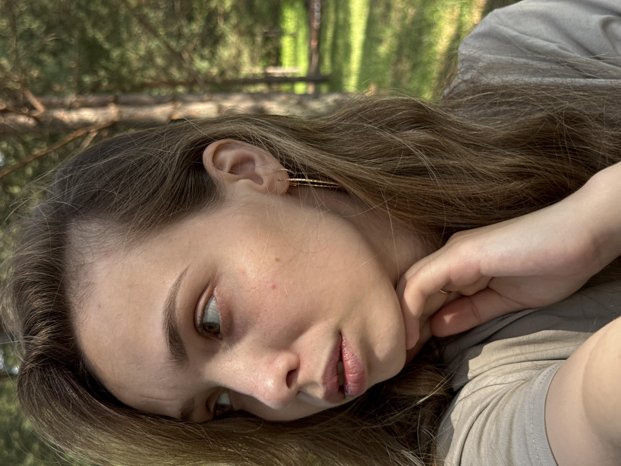 An outdoor, close-up, medium shot shows a young Caucasian woman from the chest up, looking to her left. Her long, brown hair is down and partially obscuring her right ear. She is wearing a beige t-shirt and a gold hoop earring in her left ear. Her left hand is supporting her chin. The background is blurred and appears to be a forest with trees and green grass, consistent with the location of Гольяны, Russia. The lighting suggests it is daytime with sunlight filtering through the trees.