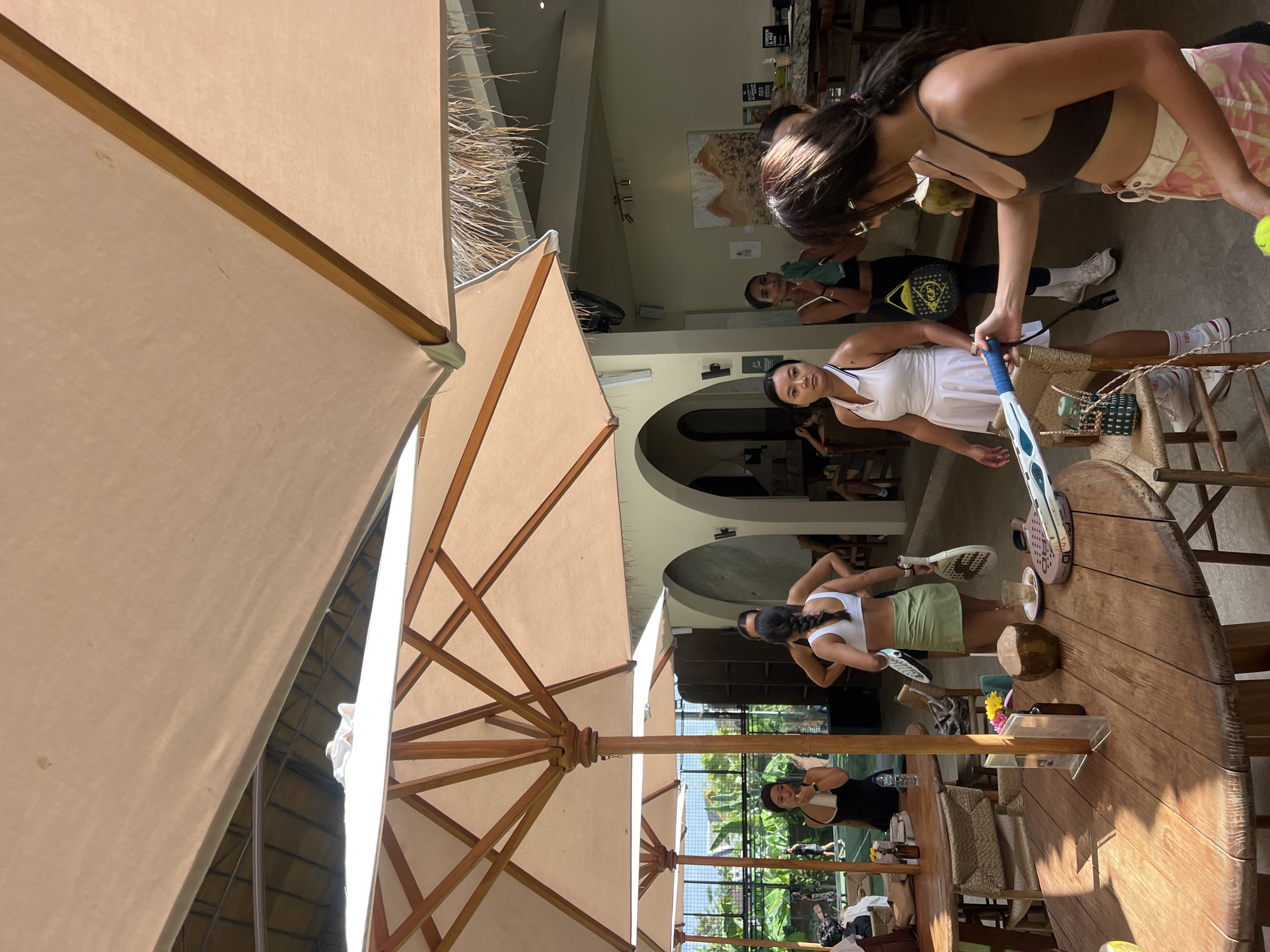 In North Kuta, Indonesia, a group of young women are gathered at an outdoor venue with large umbrellas providing shade. In the foreground, a woman with dark hair, wearing a brown bikini top and a patterned skirt, holds a tennis racket and a coconut drink. To her left, another woman in a white tennis dress stands with a tennis racket. Behind them, several other women are visible, some in athletic wear, appearing to be in a social setting. The environment suggests a relaxed, possibly sports-related gathering with elements of a cafe or resort. In the background, a tennis court and lush greenery are visible, hinting at the location's recreational purpose.