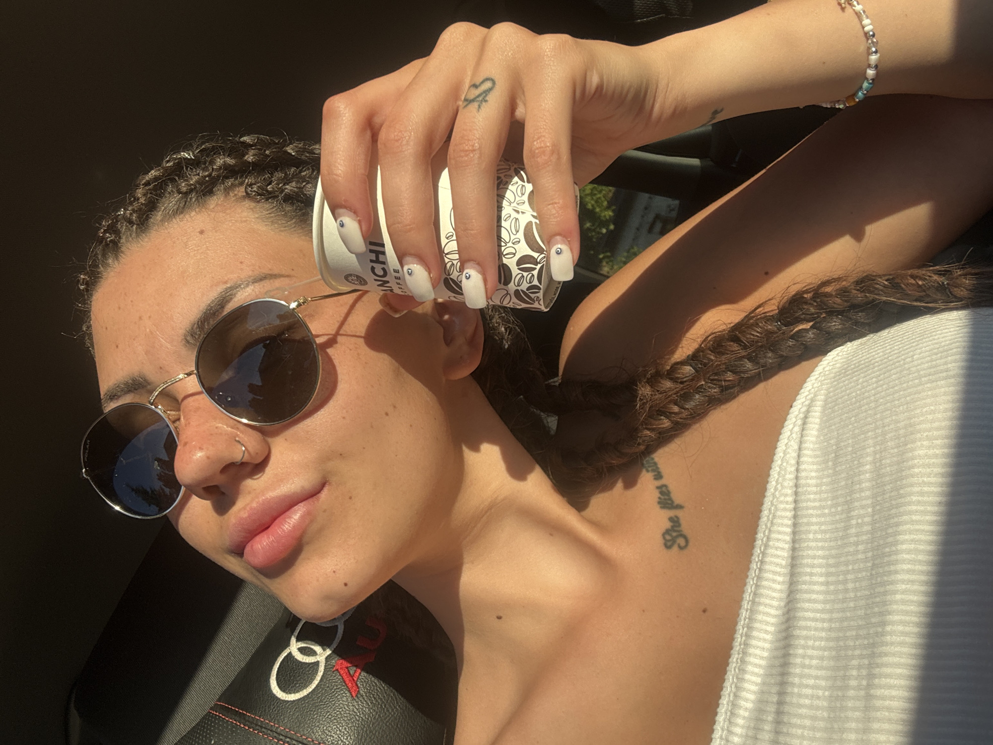 A woman with braided dark hair and a nose ring is in the driver's seat of a car, holding a coffee cup and wearing a white top and sunglasses. She is in Gorna Malina, Bulgaria, and the car interior is visible, including part of an Audi logo on the seat. The lighting suggests it is daytime, possibly late afternoon, with sunlight casting shadows. A tattoo is visible on her chest and another on her arm. She is also wearing a beaded bracelet.