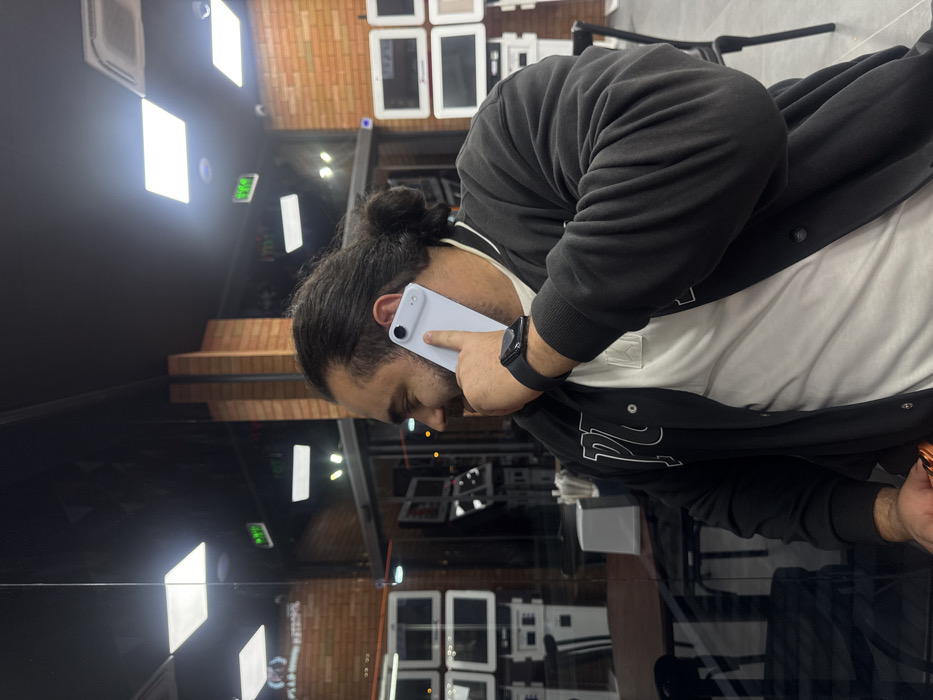 A male individual with dark hair tied in a bun, a full beard, and a mustache is depicted in a commercial indoor setting. He is wearing a dark, possibly black or dark grey, varsity-style jacket with light-colored lettering over a white t-shirt. A black smartwatch is visible on his left wrist. He holds a light grey or white smartphone to his left ear, appearing to be engaged in a phone call or listening. His expression suggests amusement or engagement, with a slight smile visible.
The scene is dominated by a prominent reflection, likely from a large glass partition or window, which forms much of the background. Through this reflection, the interior of a modern establishment is visible, characterized by dark ceilings with square recessed LED lighting fixtures and sections of exposed brick walls. Multiple white-framed digital displays or monitors are arranged in grids on the walls, suggesting a retail or service environment such as a cafe, fast-food outlet, or electronics store. A green illuminated exit sign is also discernible in the reflection. A dark table with a white napkin dispenser and dark chairs are visible, indicating a seating area. The specific location in the city of Yerevan, Armenia, is not identifiable from the visual information provided.