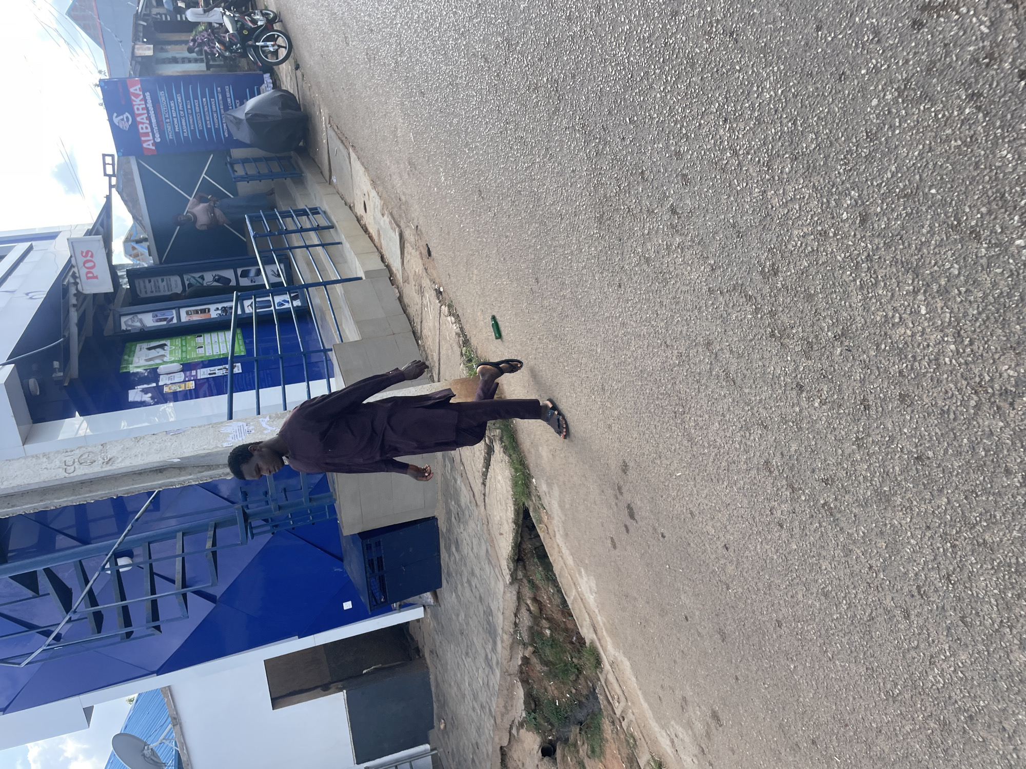 A man in a dark purple traditional outfit walks on a paved road in Jos, Nigeria. He is carrying a brown paper bag in his right hand and wearing black sandals. The road is a light gray asphalt with some patches and small stones. To his left, there is a gutter with some water and greenery. Behind the gutter, there is a sidewalk with a white and blue tiled building. The building has an external metal staircase on its left side, painted blue. Further to the right, there is a shop with a "POS" sign and another shop with a large blue and white sign that reads "ALBARKA Communications". A motorcycle is parked on the right side of the road, and a woman stands in the doorway of a building to the right of the Albarka Communications sign. The sky above is partly cloudy.