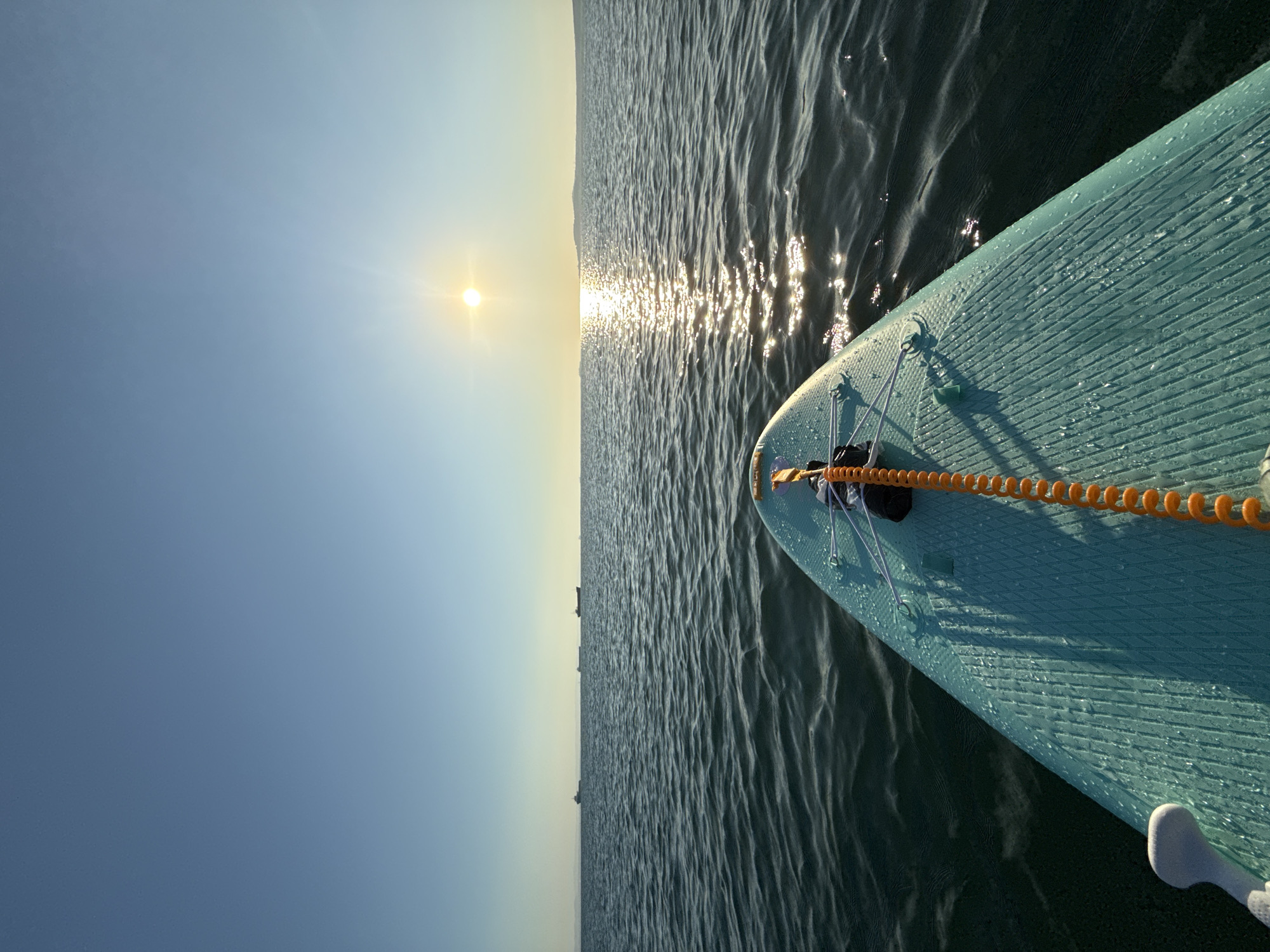A point-of-view shot from a stand-up paddleboard on the sea. The paddleboard, colored a light teal and covered in water droplets, dominates the foreground. An orange coiled leash extends from the board's nose towards the viewer, secured by a black buckle. The sea is a deep blue-green with gentle waves and is illuminated by the sun, creating a shimmering reflection that stretches towards the horizon. In the distance, several cargo ships are visible on the water, and a faint landmass can be seen on the far horizon. The sky is a clear, pale blue with the sun positioned in the upper right quadrant. The scene appears to be set in the Burgas Bay, near the city of Burgas, Bulgaria, as indicated by the presence of cargo ships commonly seen in the area and the general seascape. There are no people visible in the frame.