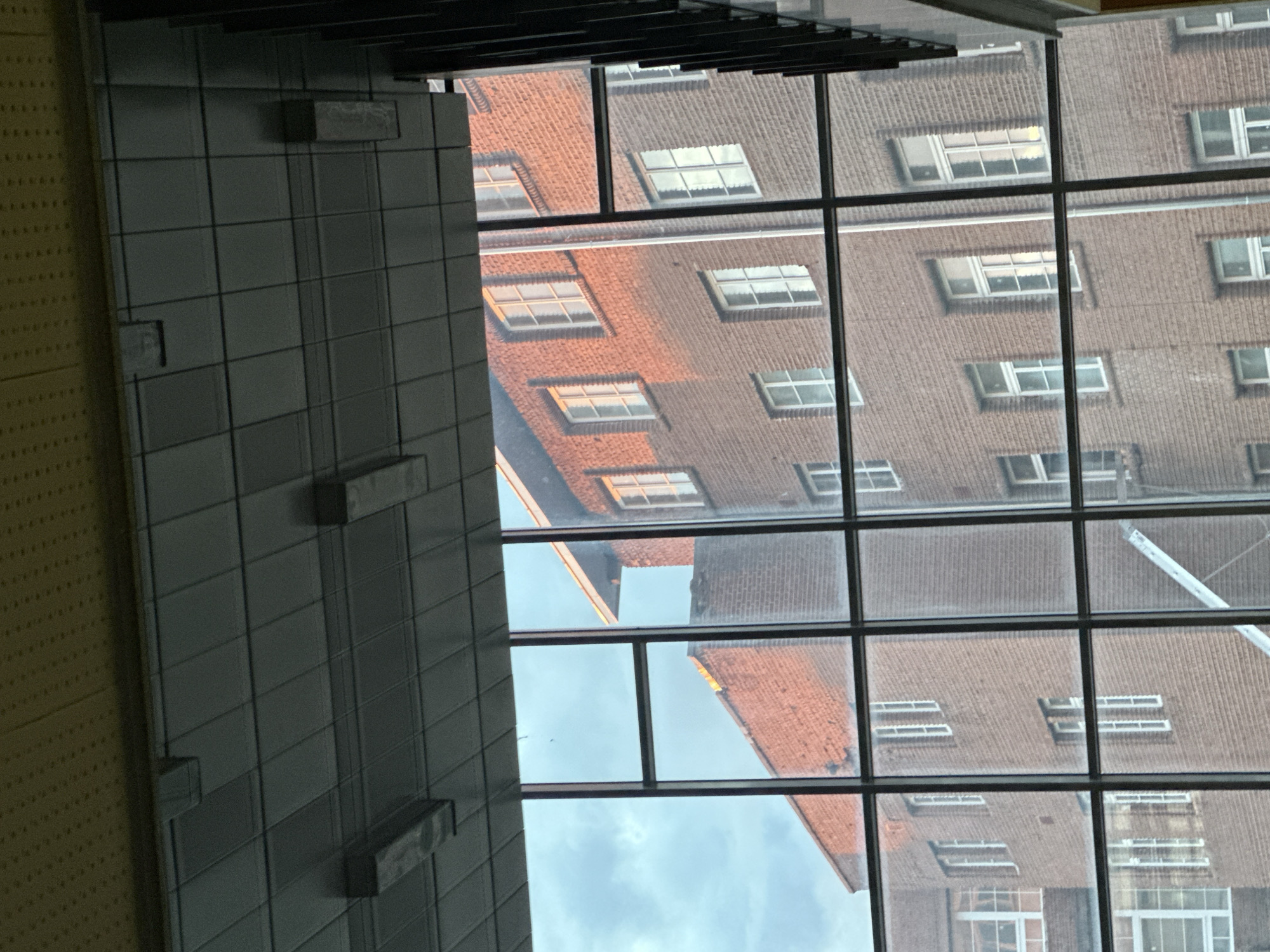 The image shows a view through a multi-pane window. Reflected in the window are brick buildings with multiple windows. Portions of the sky are visible through some panes. A grey tiled wall is partially visible on the left side of the frame. A black ventilation system is visible at the top of the frame. The image might be taken indoors, looking out at the city of Sofia, Bulgaria.