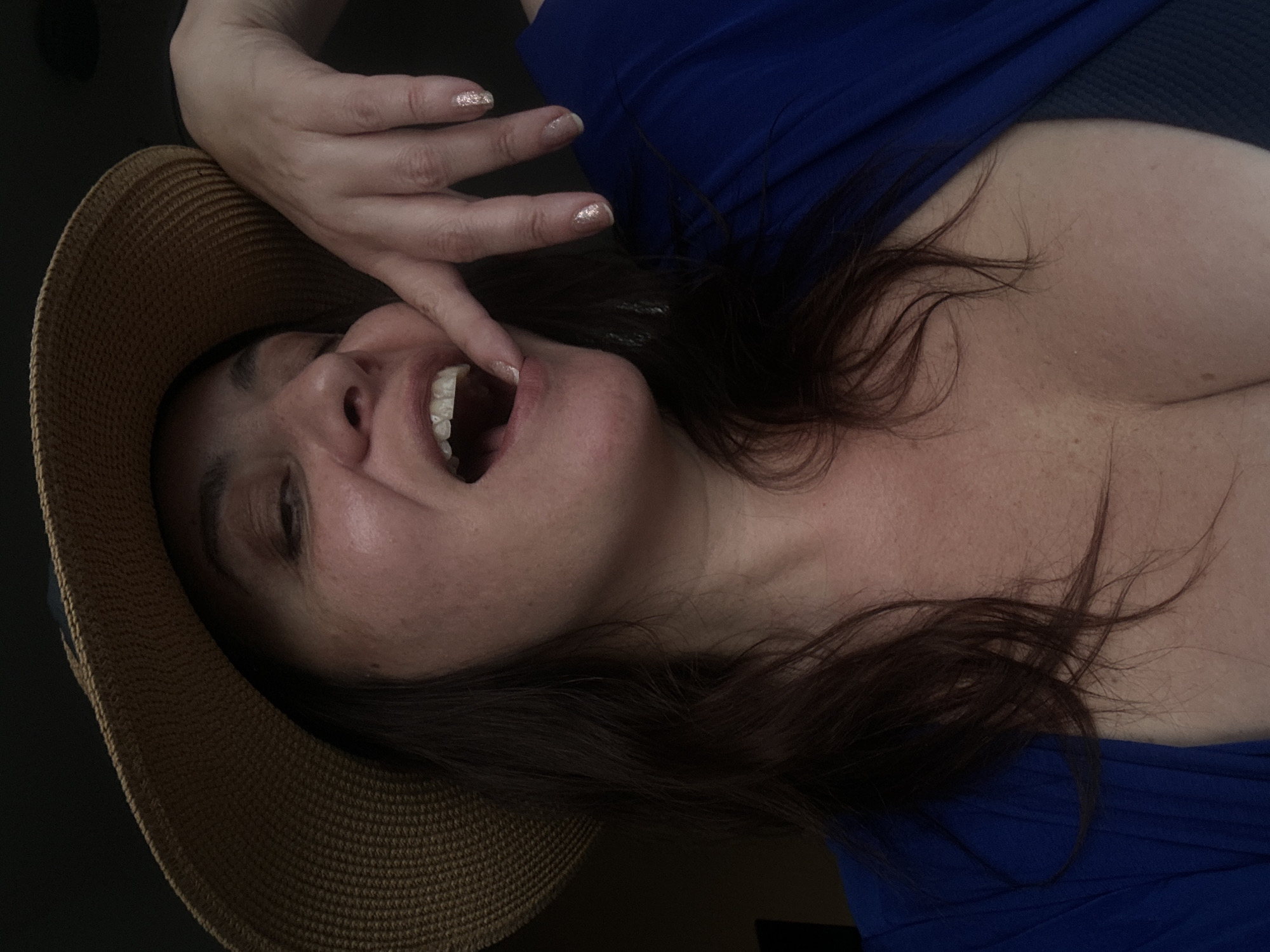 The image shows a close-up of a woman wearing a straw hat and a blue top. She has her mouth open and her finger is touching her lips. Her nails are decorated with glitter. The background is dark and indistinct. The image does not provide any information about the location, including whether it is in Riga, Latvia.