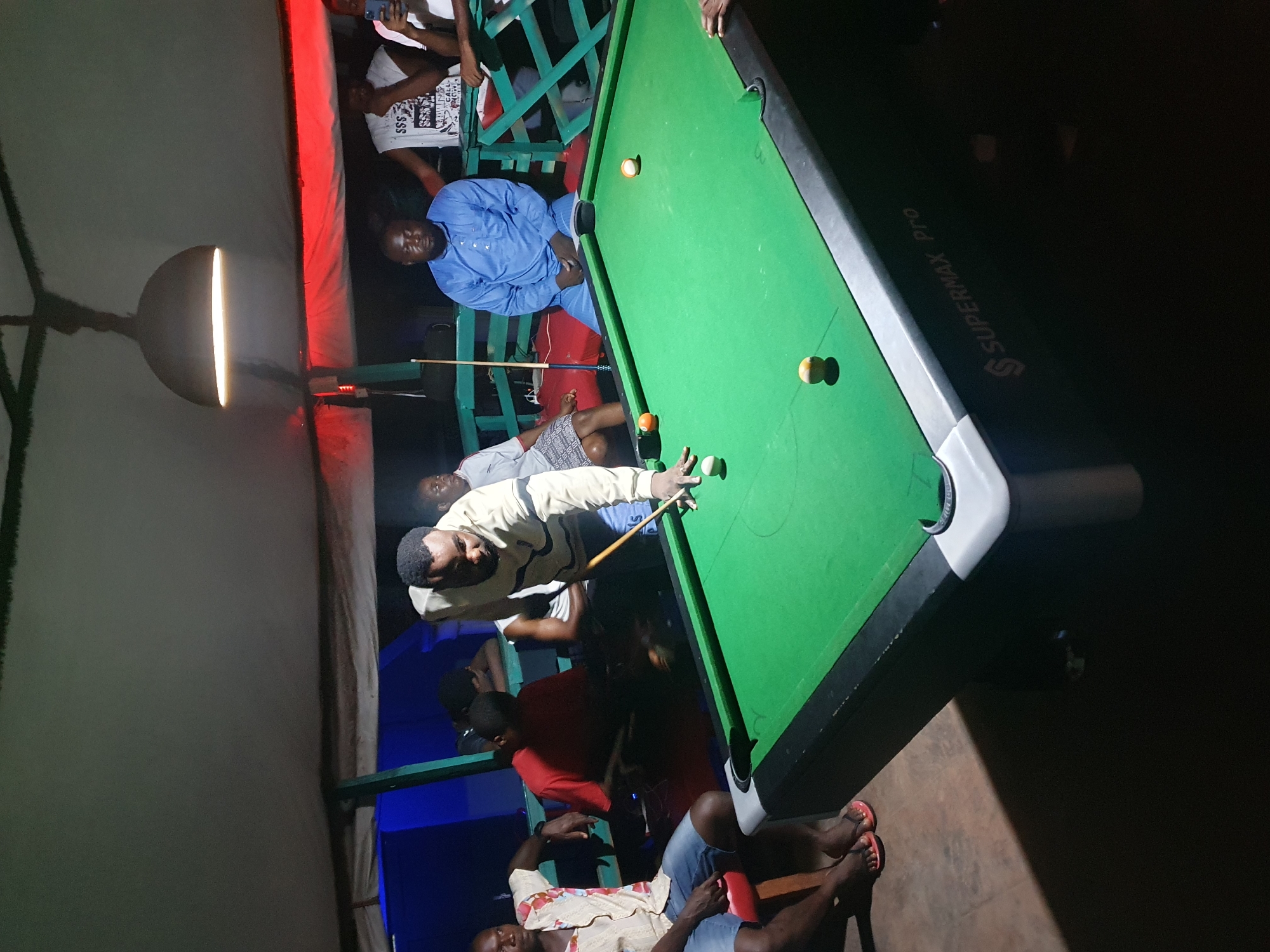 A group of people are gathered around a pool table in a dimly lit space. The pool table is green with several balls on it. One man is taking a shot. People are seated around the table, watching the game. The environment appears to be a casual entertainment venue, possibly a bar or club. The location is Lokoja, Nigeria.