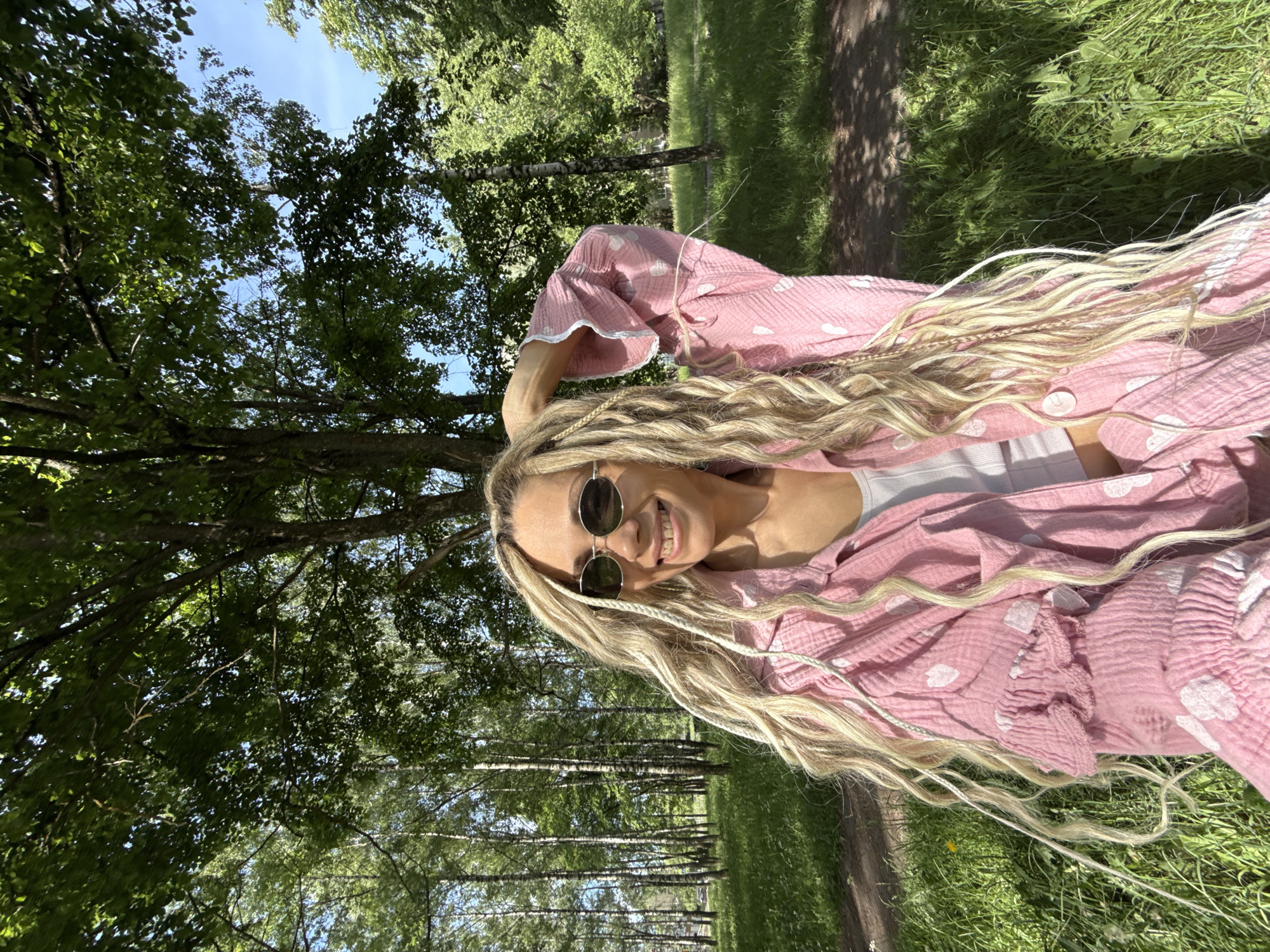 A female subject is positioned outdoors in a park-like setting under clear blue sky, illuminated by bright daylight. The subject, with fair skin and long, light blonde hair styled with a combination of dreadlocks, braids, and wavy sections, some accented with white thread, is wearing dark, oval-shaped sunglasses with silver frames. She is smiling broadly, revealing teeth. Her left hand is raised to the back of her head, lightly touching her hair. She is dressed in a light pink, textured fabric shirt or jacket patterned with white heart shapes, featuring ruffled detailing on the sleeves and a visible white crop top underneath. Matching pink bottoms with white heart patterns and a ruffled hem are also visible.
The background consists of a dense green environment. To the left, a grove of slender birch trees with white bark is visible amidst green foliage, with a faint indication of urban structures or a road in the distant background. To the right and overhead, large trees with full green canopies obscure parts of the sky. The ground is covered in green grass with patches of earth or pathways and some weeds. Shadows from trees are cast upon the ground, indicating direct sunlight. The scene is located in the city of Unknown, Unknown.