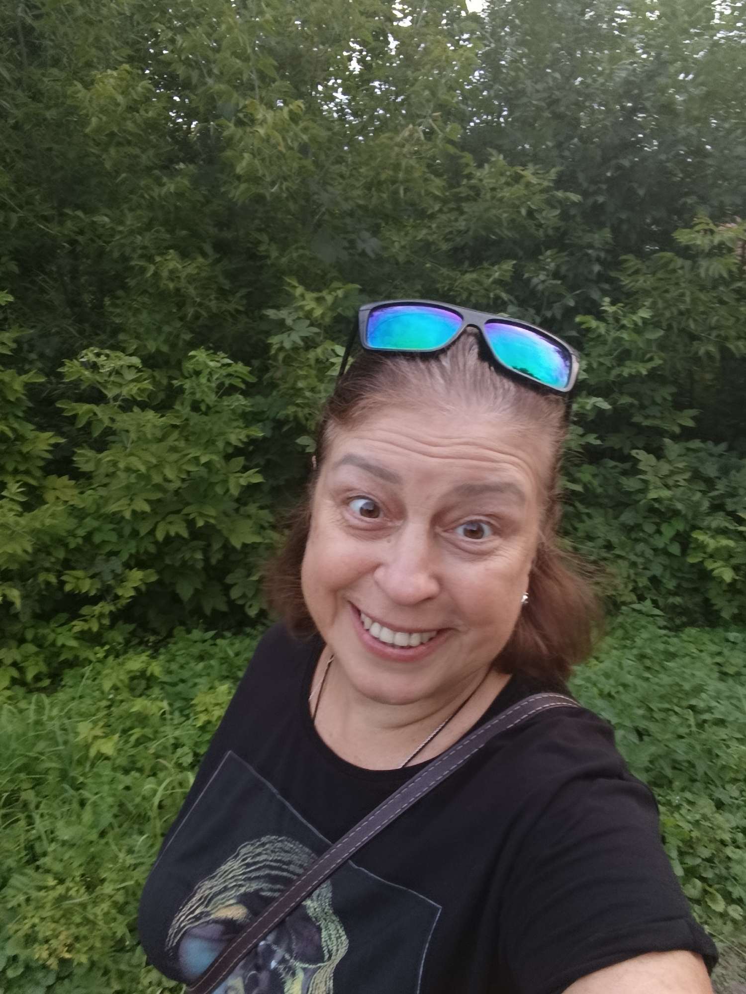 A woman with brown hair, wearing sunglasses, and a black t-shirt stands in front of a dense green foliage background. She is smiling broadly at the camera, with her mouth open and teeth visible. The sunglasses have iridescent lenses that reflect blue and green. She is wearing a simple necklace, and a strap of a bag is visible crossing her chest. The overall setting appears to be outdoors, possibly a park or wooded area. There is no specific indication of the city of Vereya, Russia, within the image itself.