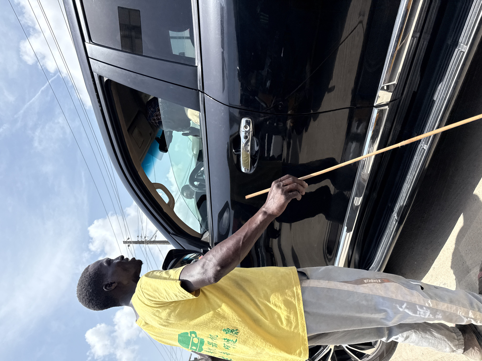 A man in Lagos, Nigeria, stands beside a black car with its door ajar. He is holding a wooden stick in his right hand, pointing towards the car. The man is wearing a yellow t-shirt and gray pants. The sky is visible with scattered clouds, and there are power lines overhead. The car is parked on a paved surface, likely a street.