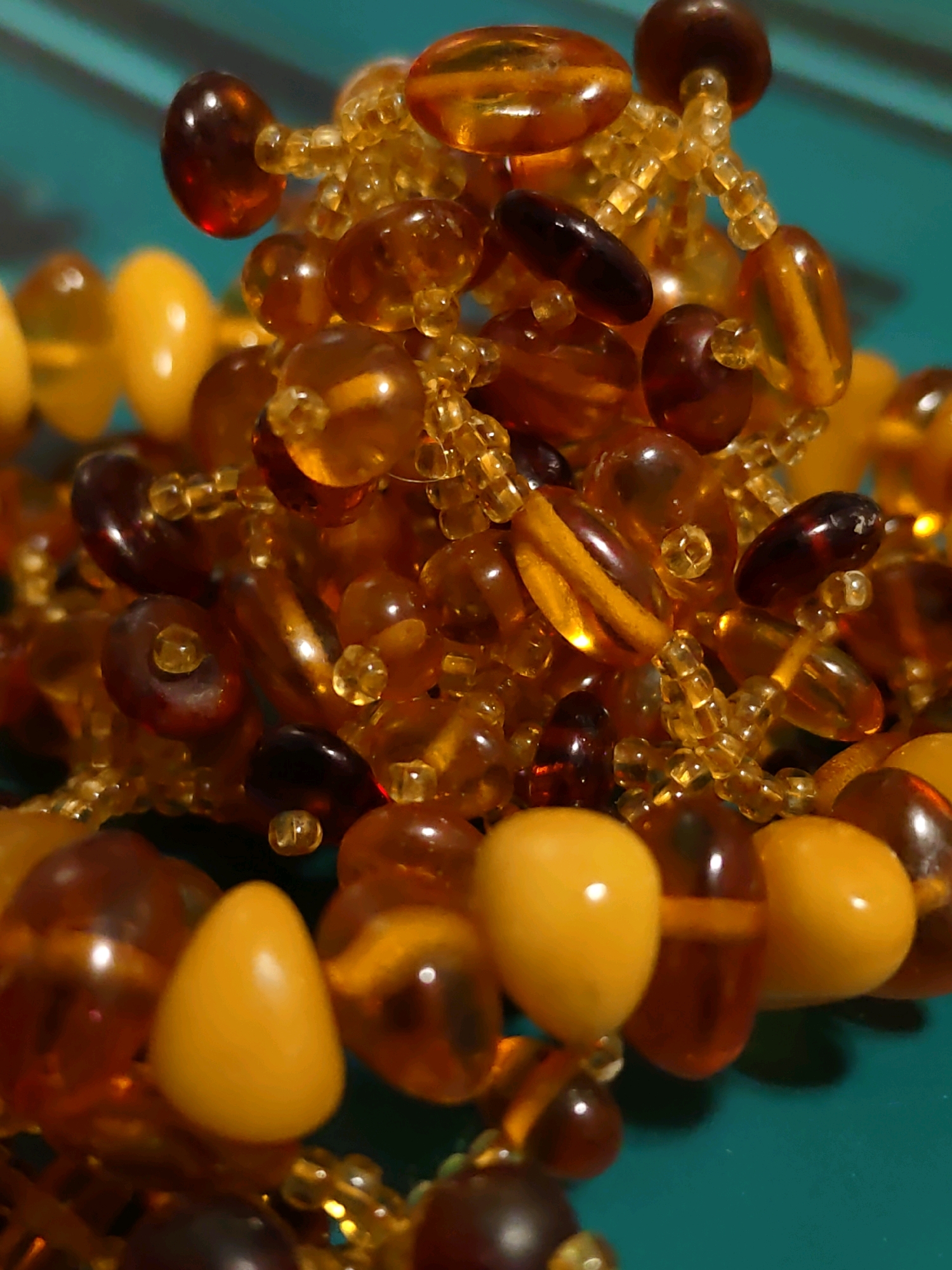 A macro photograph shows a close-up of a beaded necklace or bracelet made of amber beads and small golden seed beads. The amber beads vary in color from light yellow to deep brown, and are shaped into ovals, rounds, and teardrops. The necklace is spread across a teal-colored surface. There are no people or any discernable city elements visible in the image.