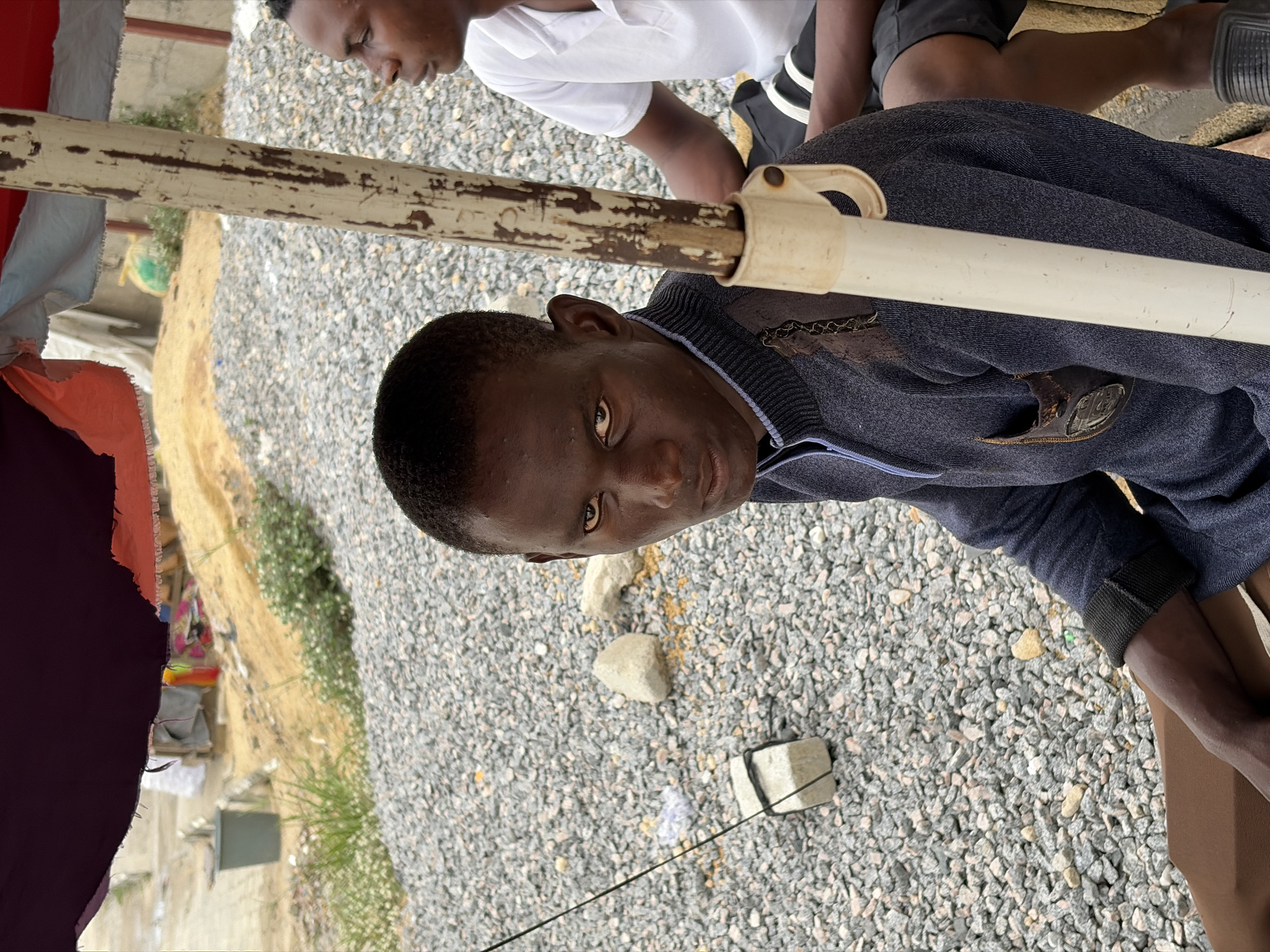A close-up shot, taken from a high angle, shows two dark-skinned men on a gravelly surface likely in Igboefon, Nigeria. The nearest man, wearing a navy zipped sweater with a leather patch, gazes directly at the camera. A worn, wooden pole with peeling paint is positioned horizontally across the image. In the background, another man is partially visible, wearing a white T-shirt. The ground is covered with small gray stones. A section of fabric, likely from a shelter, is present to the left. In the periphery, glimpses of grass and distant buildings suggest an outdoor setting.